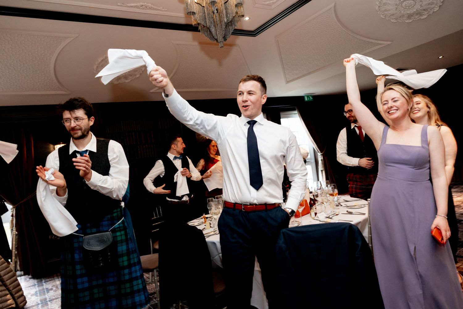 Wedding guests in kilts and formal wear wave papers and cheer during a lively reception celebration.