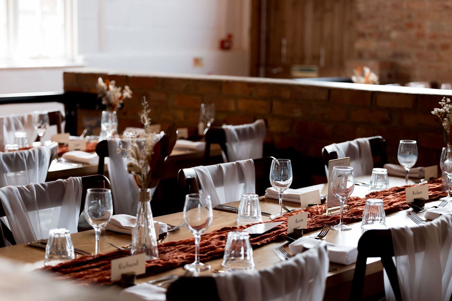 Long banquet table setup with elegant place settings against exposed brick wall.