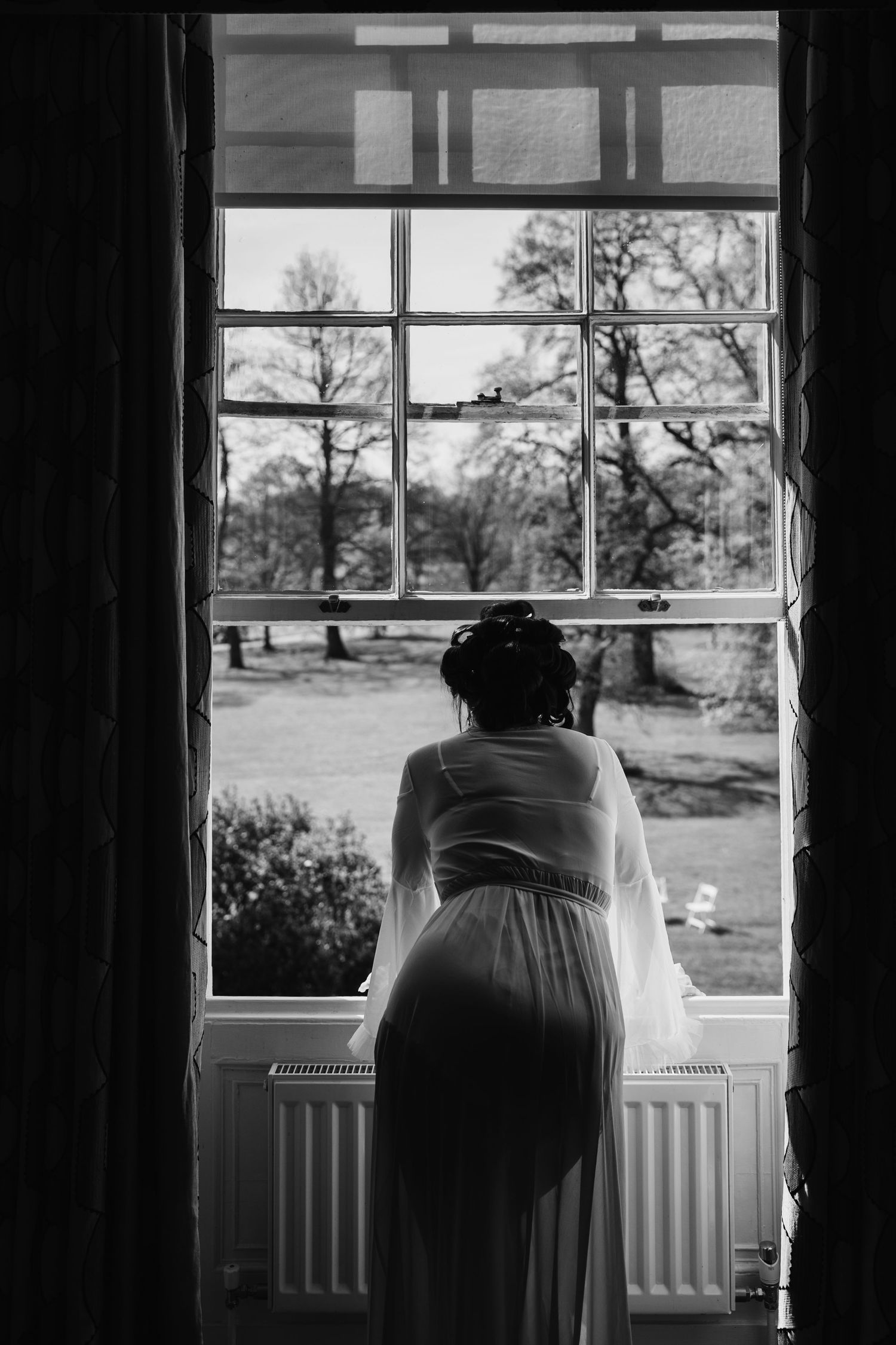 Black and white photo of a bride in a wedding dress standing at a large window overlooking a garden.