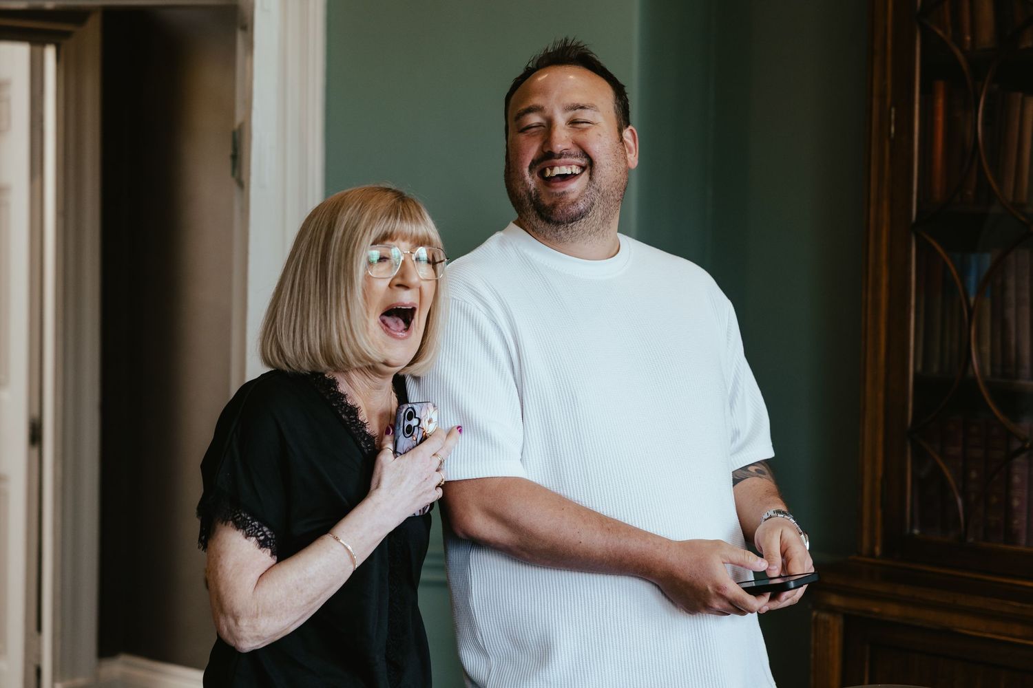 Two people laughing joyfully together indoors, woman in black lace top and man in white t-shirt sharing a fun moment.