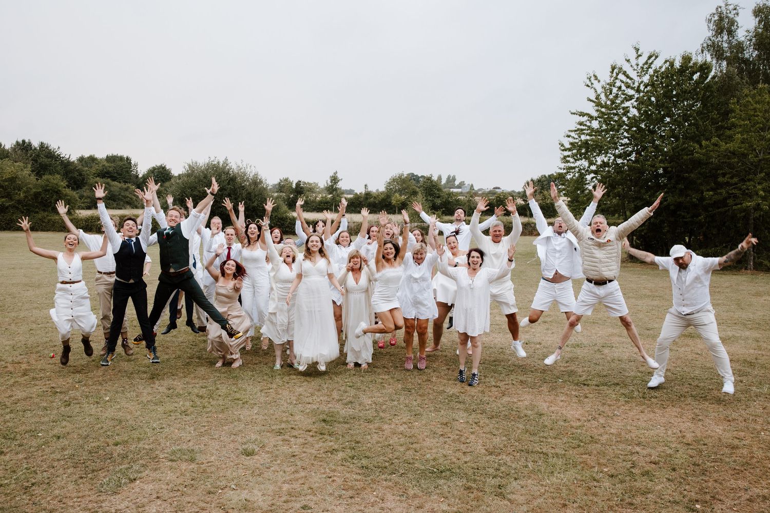 A large wedding party dressed in all white poses playfully on a grassy field.
