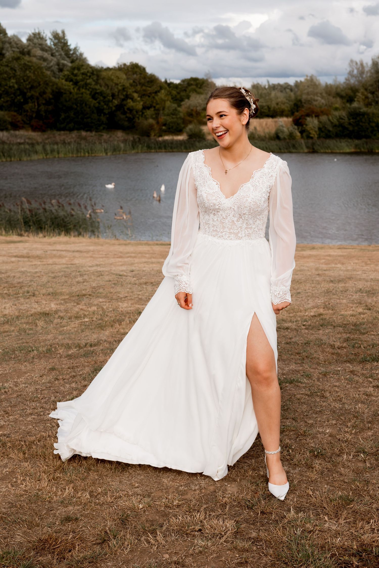 Elegant wedding dress with long sheer sleeves and thigh-high slit photographed by a lake with a swan in the background.