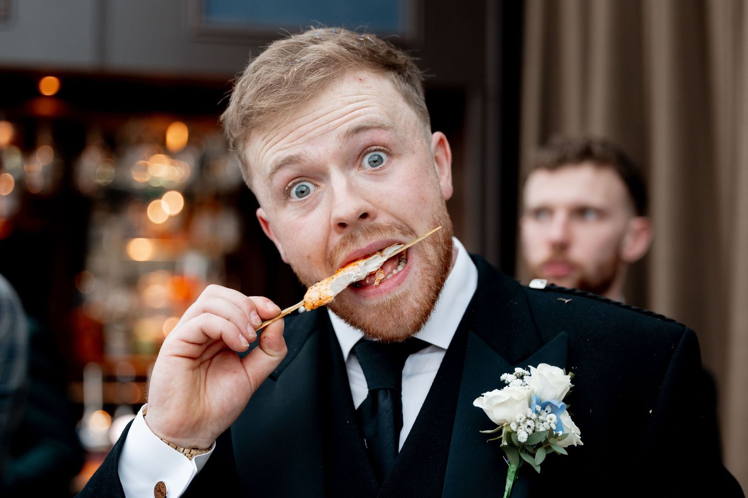Groom in black suit with boutonniere humorously biting a skewer at a wedding reception.
