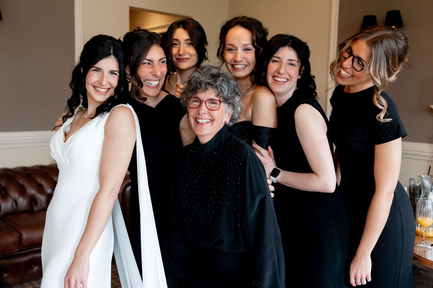 Bride in white gown smiles with bridesmaids and older woman in black dresses during wedding preparations.