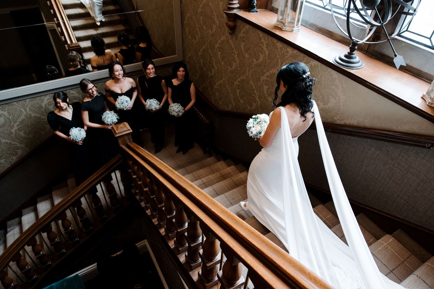 Bride in white gown descends staircase while bridesmaids in black dresses holding white bouquets watch from above.