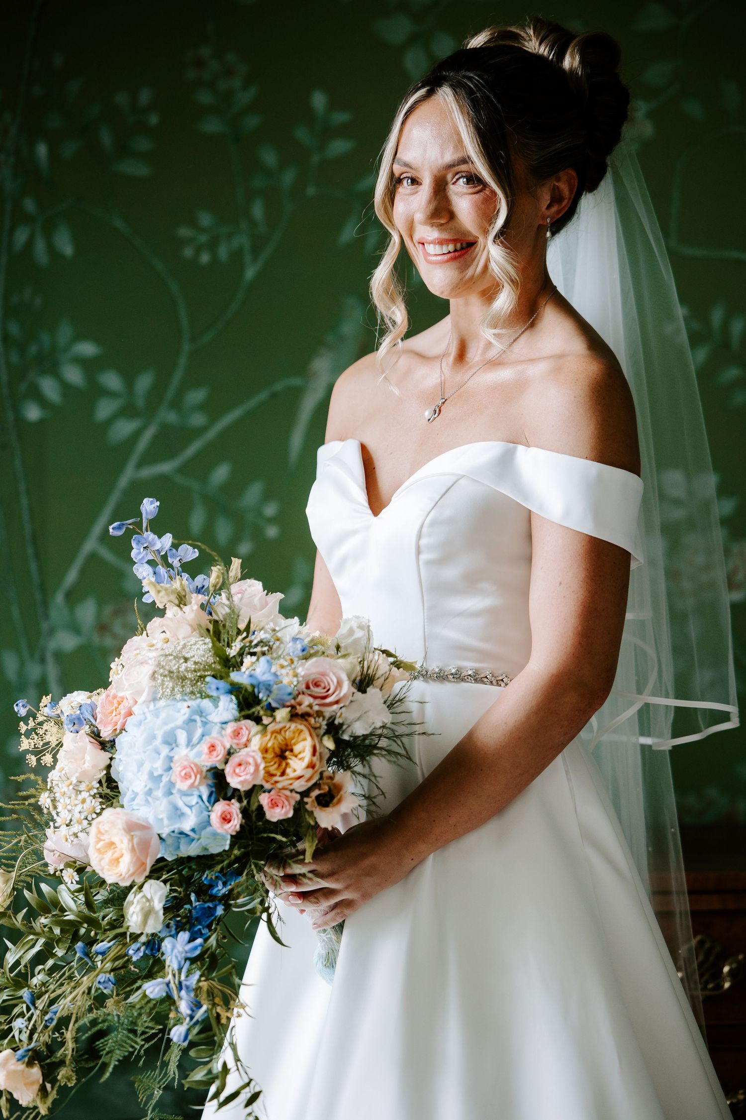 Bride in white off-shoulder gown holds lush pastel bouquet against green foliage backdrop.