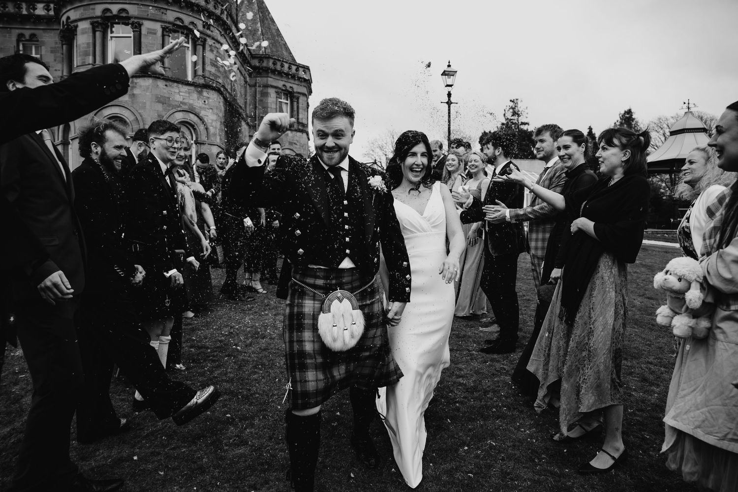 Groom in Scottish kilt walks with bride after wedding ceremony, surrounded by cheering guests.