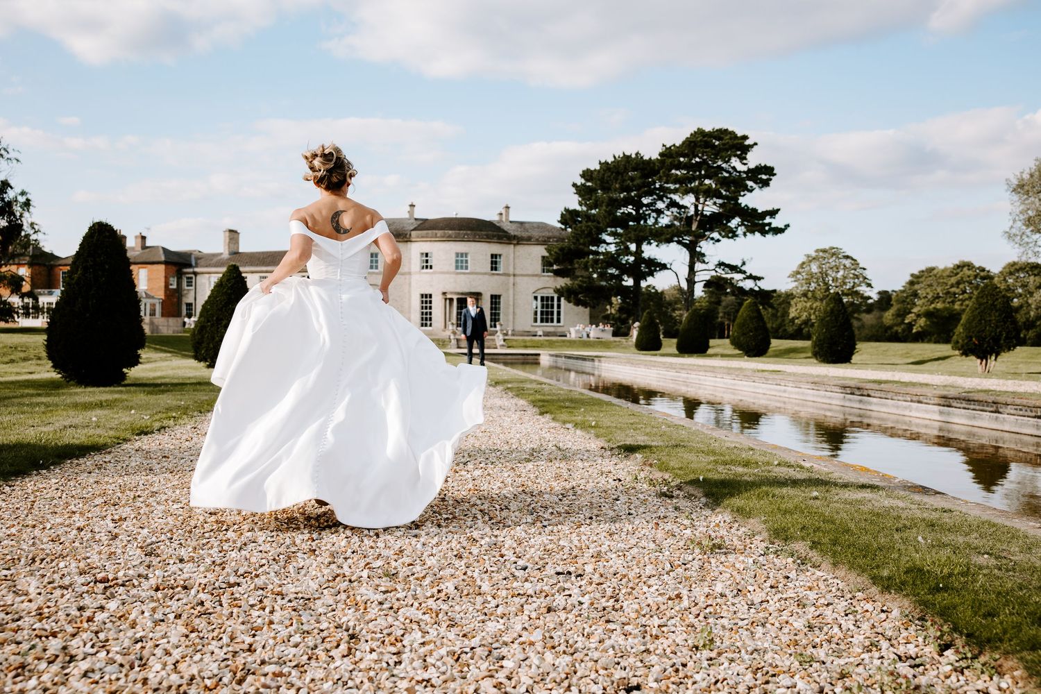 A white wedding dress flows elegantly across a gravel path beside a tranquil pond on a sunny day.