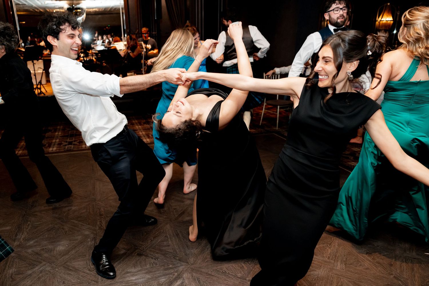 Guests dance energetically at a wedding reception, with women in black and green gowns spinning on the floor.