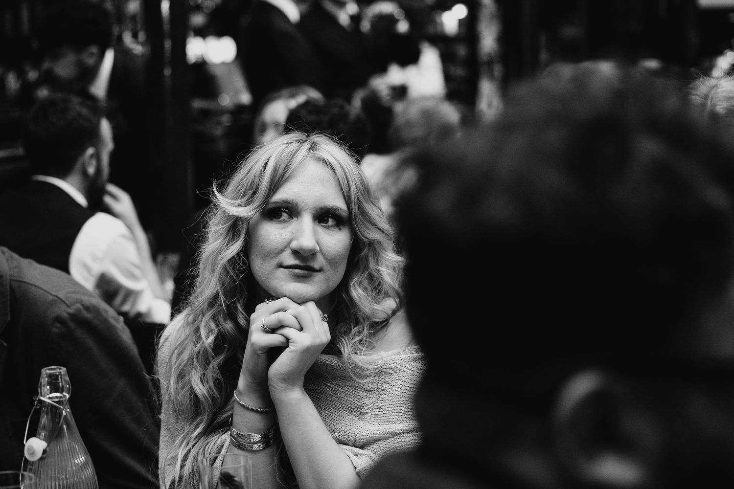 Black and white photo of a smiling woman with wavy blonde hair sitting at a social gathering, hands clasped near her chin.