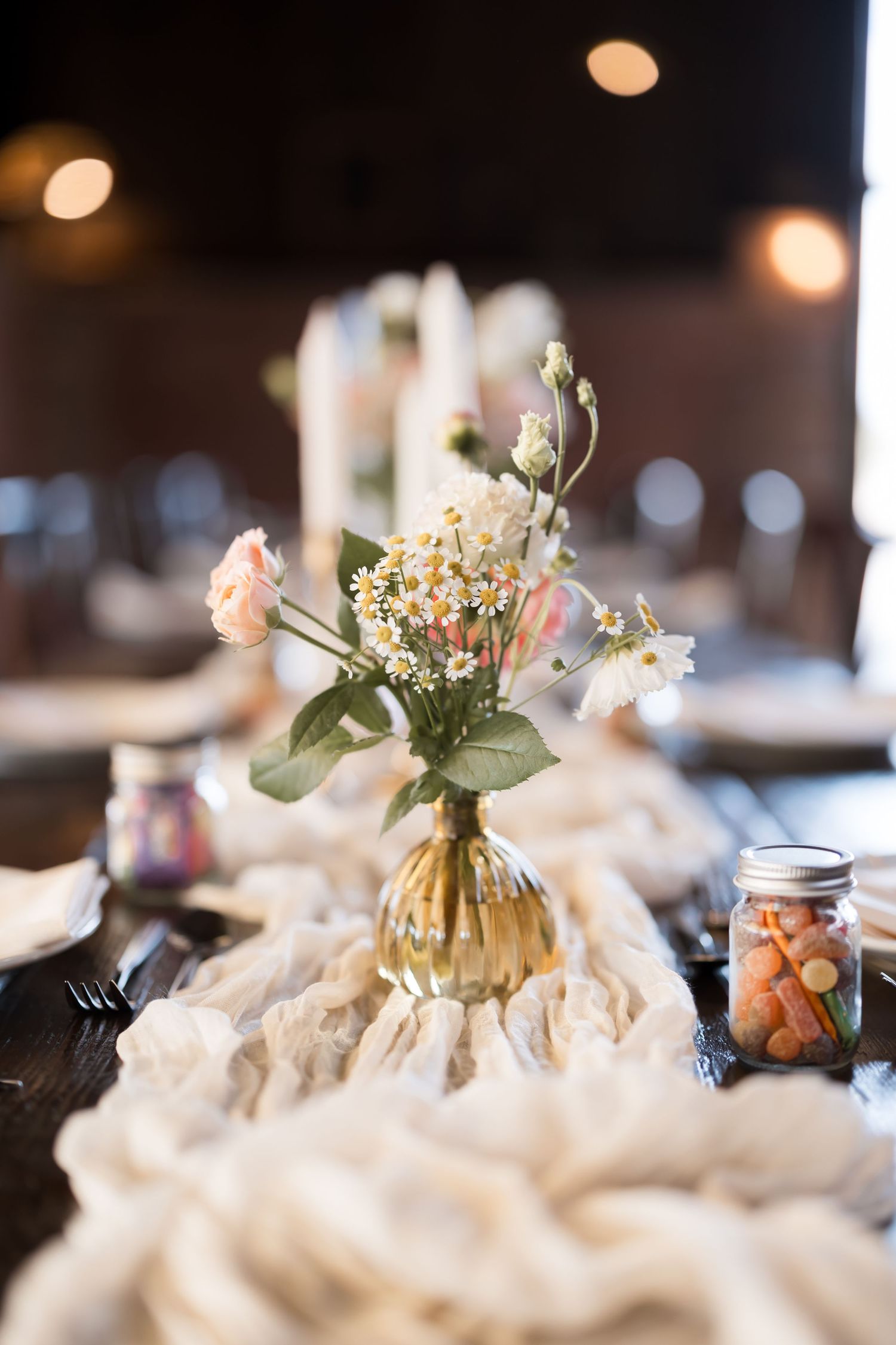 Elegant wedding table centerpiece with pink roses and daisies in a glass vase on a white gauze runner.