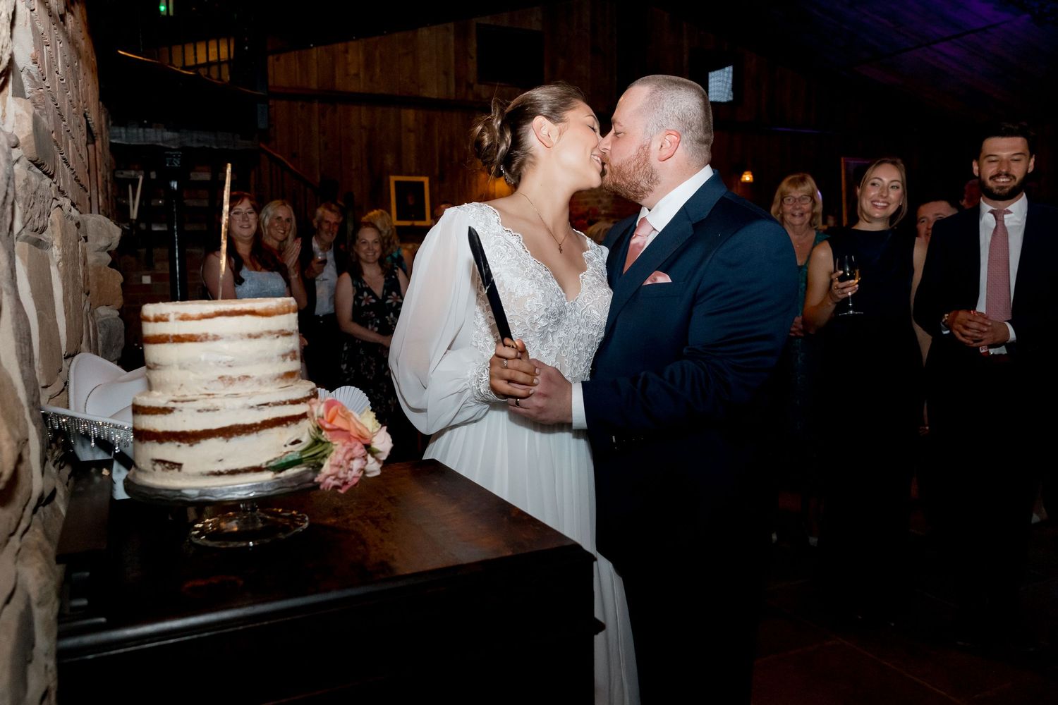 Wedding couple sharing a kiss next to their semi-naked wedding cake at a rustic reception.