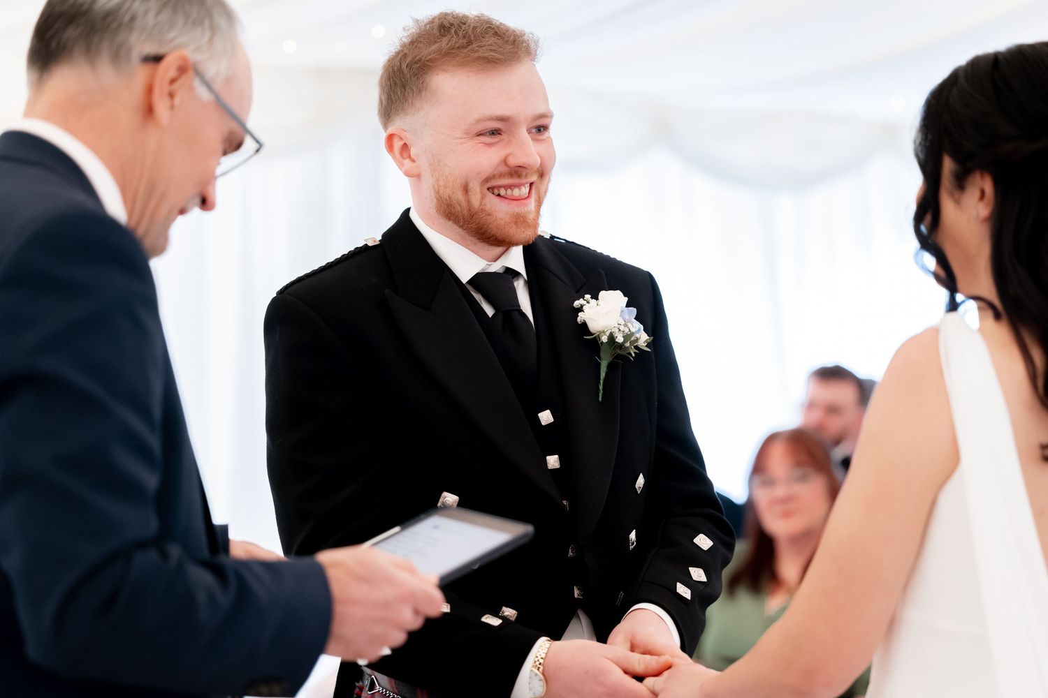 Smiling groom in Scottish kilt holding hands with bride during wedding ceremony.