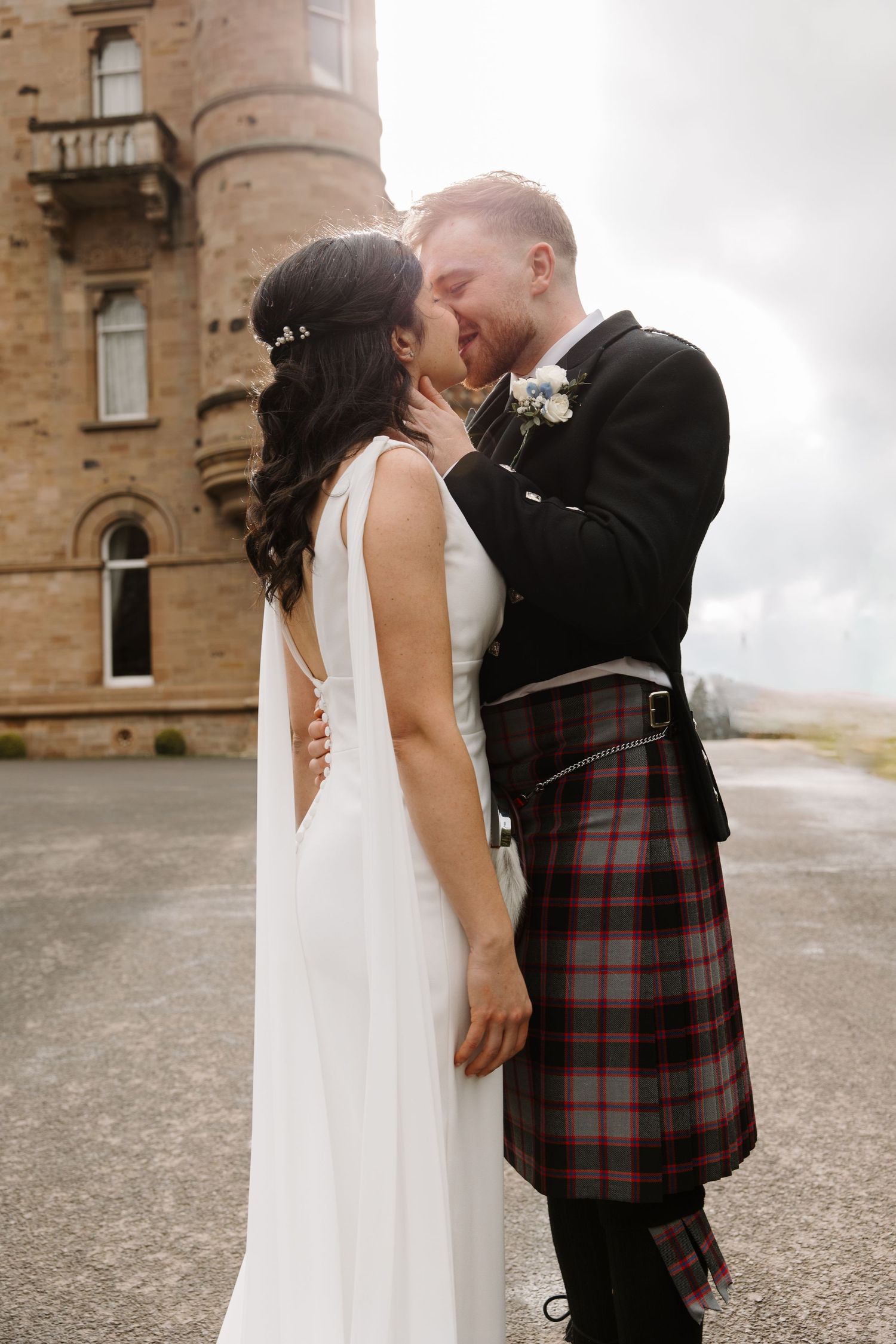 Bride and groom sharing a kiss outside a Scottish castle, groom wearing a traditional tartan kilt.