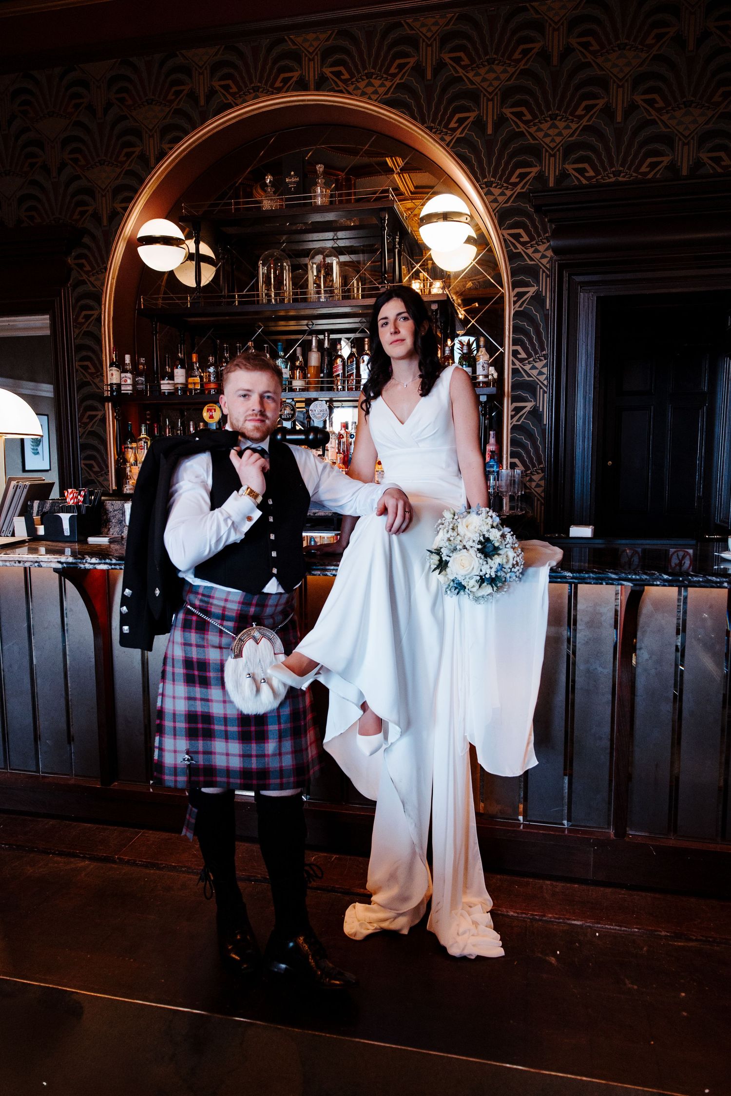Bride and groom posing at a dark ornate bar, groom in tartan kilt holding a drink.