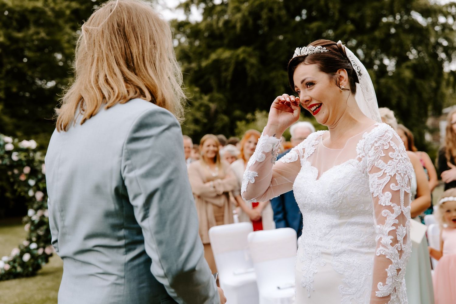 Emotional bride in lace gown wipes tear during outdoor wedding ceremony while guest in sage suit looks on.