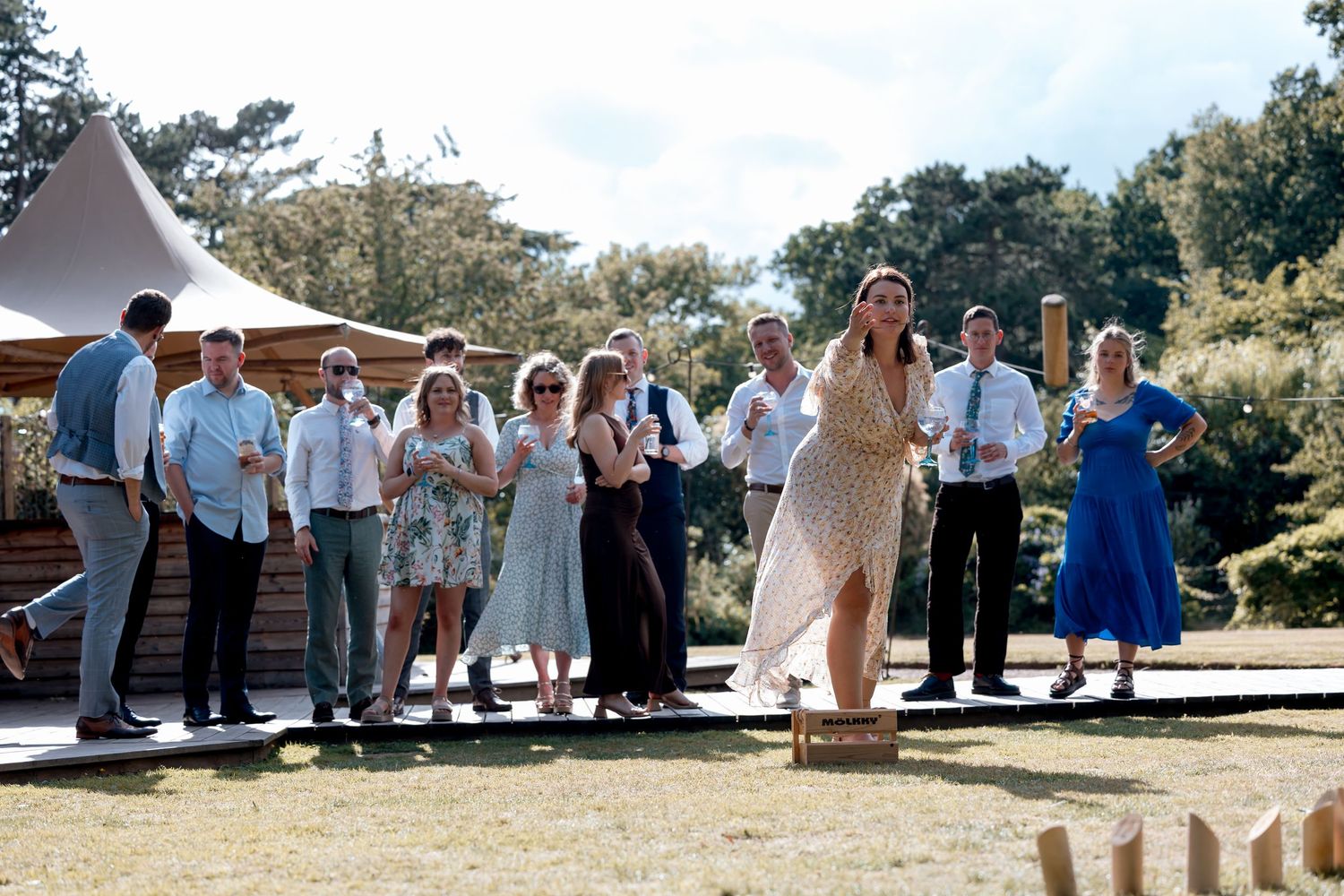 Wedding guests gather under white tent canopies on a sunny outdoor lawn.