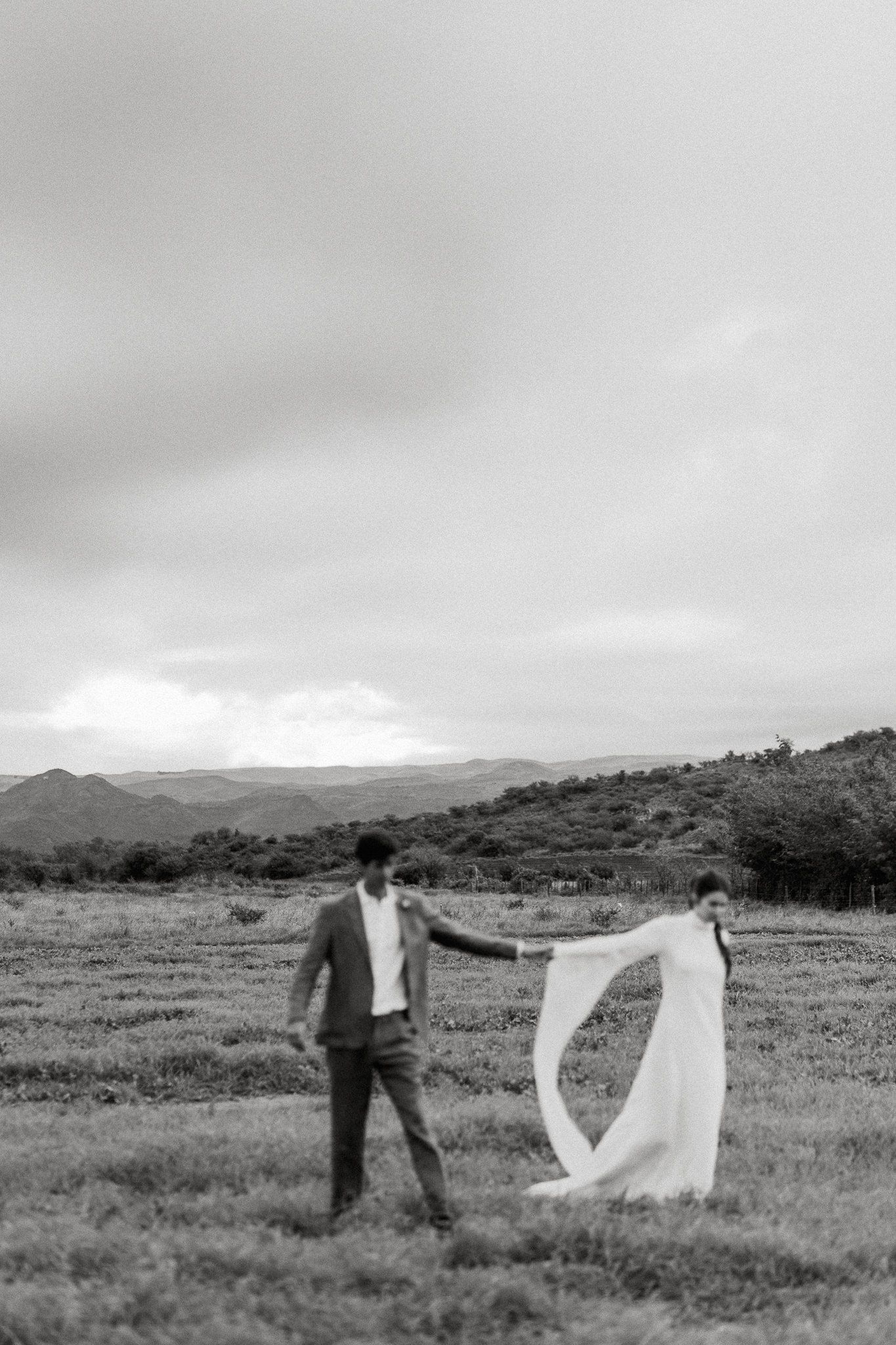 A couple in wedding attire holds hands in a scenic mountain landscape on an overcast day, captured in black and white.