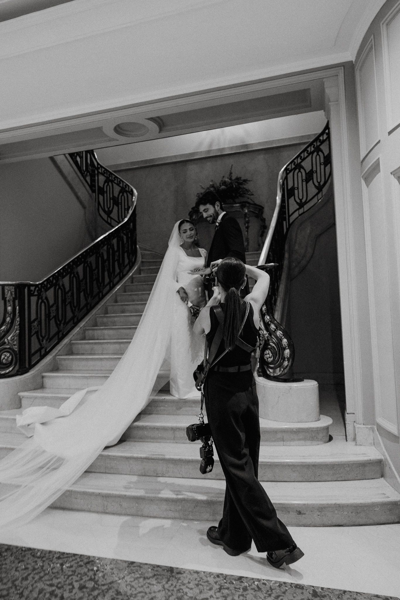 A flowing white wedding gown trails down an elegant marble staircase with ornate black iron railings in a black and white photo.