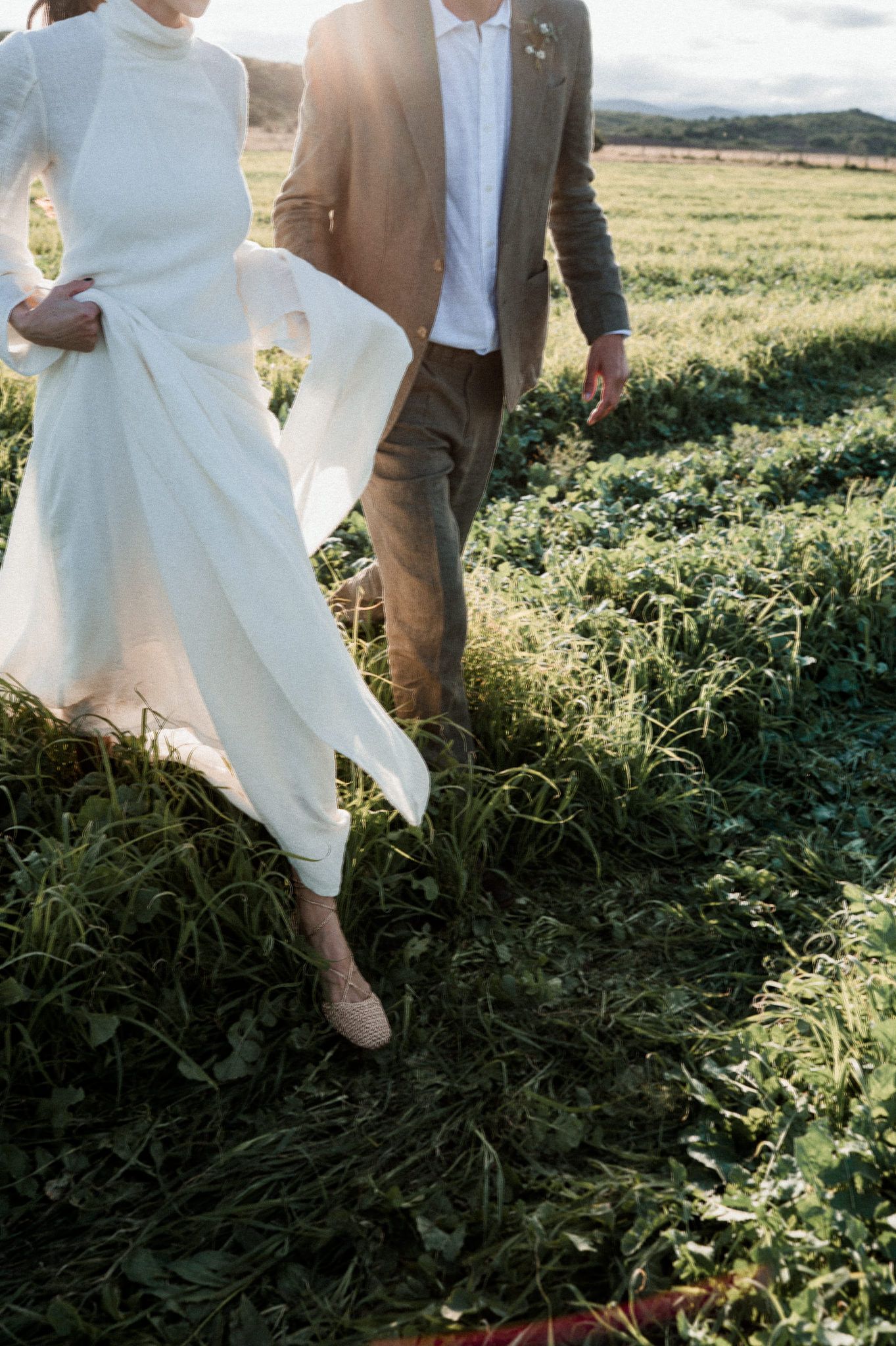 A couple in wedding attire walks through a sunlit field with tall grass and wildflowers on a bright summer day.