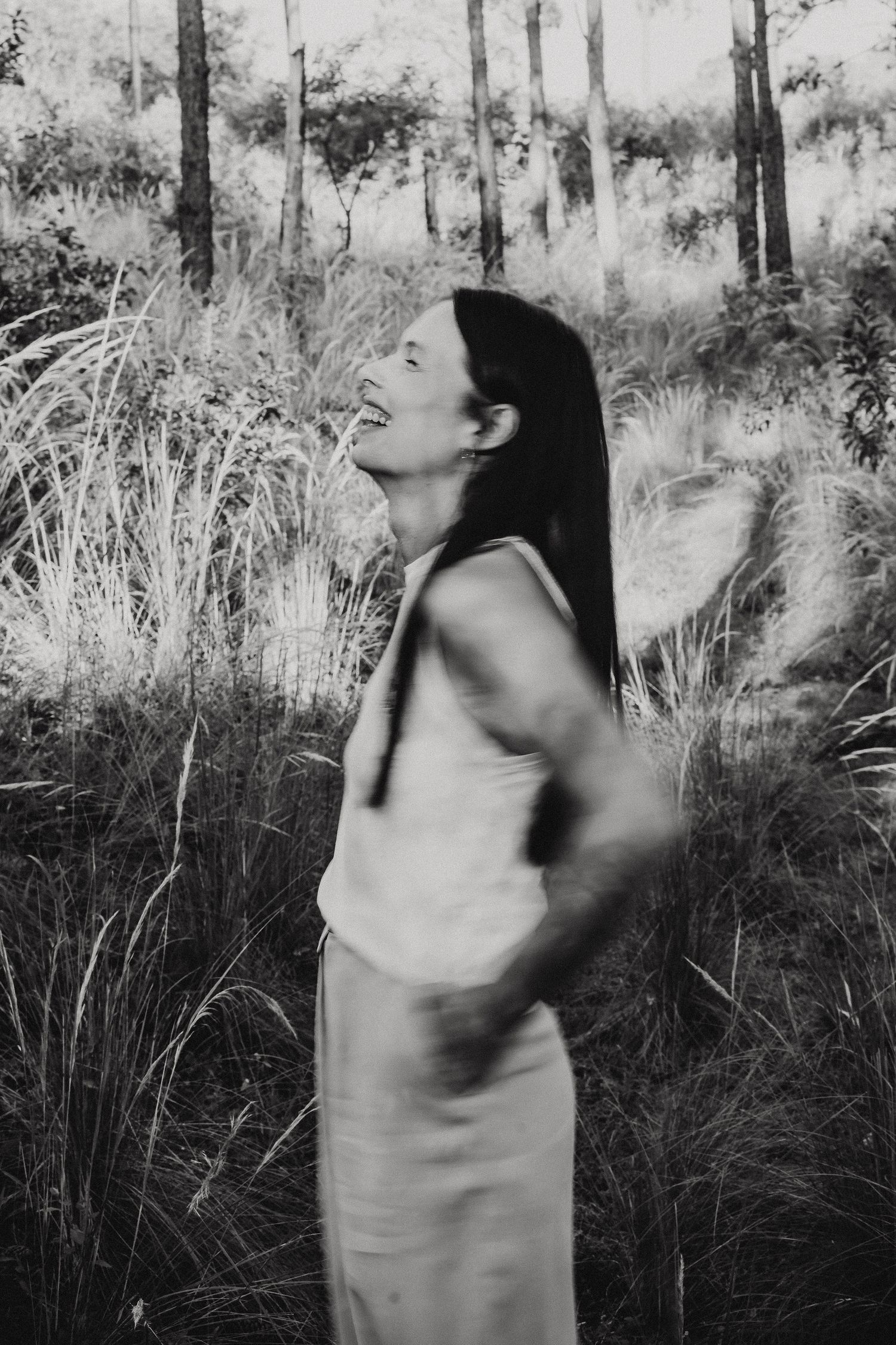 Black and white artistic photograph of a person in a light dress standing among tall grass and trees in a forest setting.