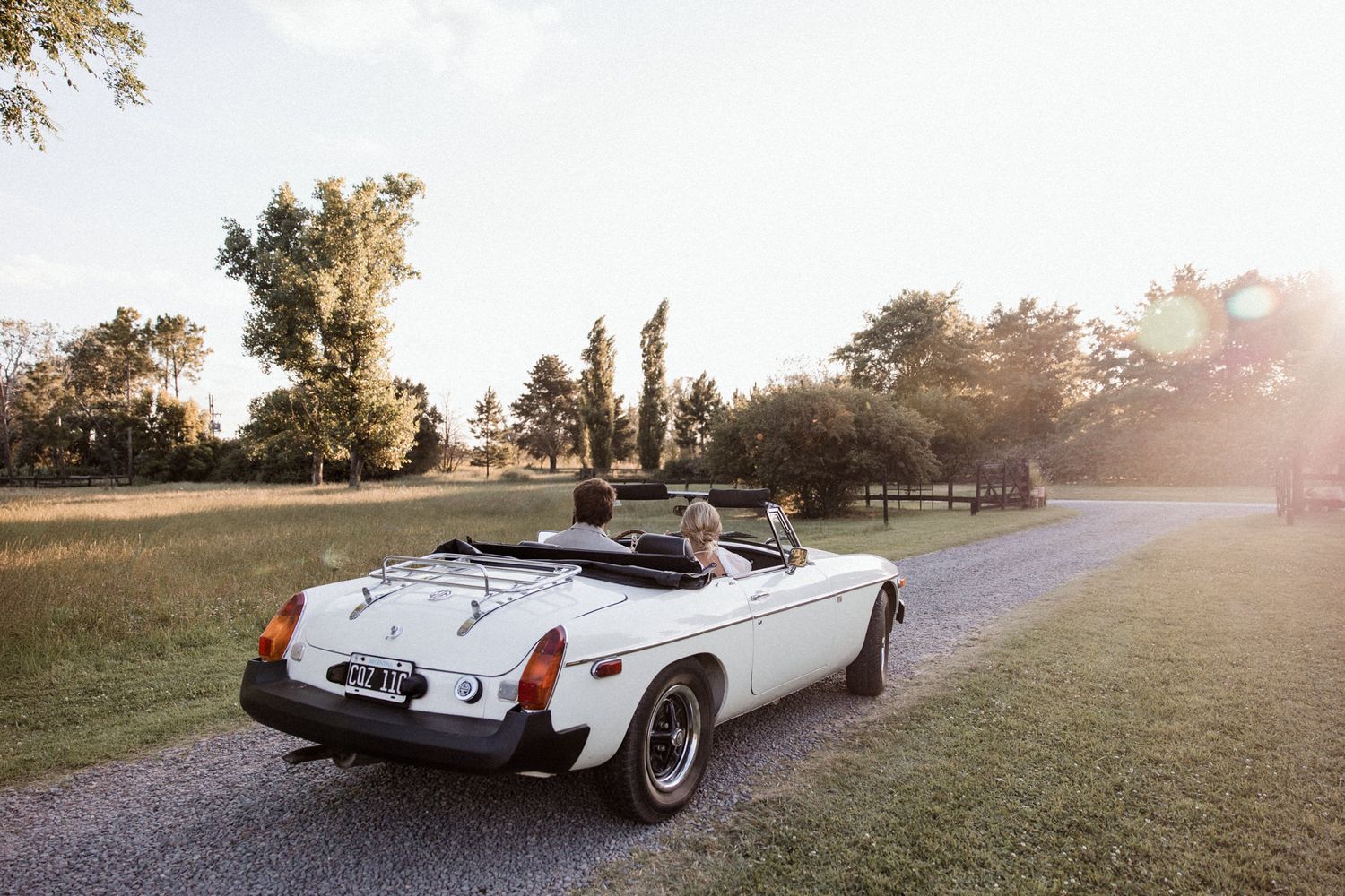 Couple riding in a vintage white MGB convertible sports car down a gravel path at golden hour.