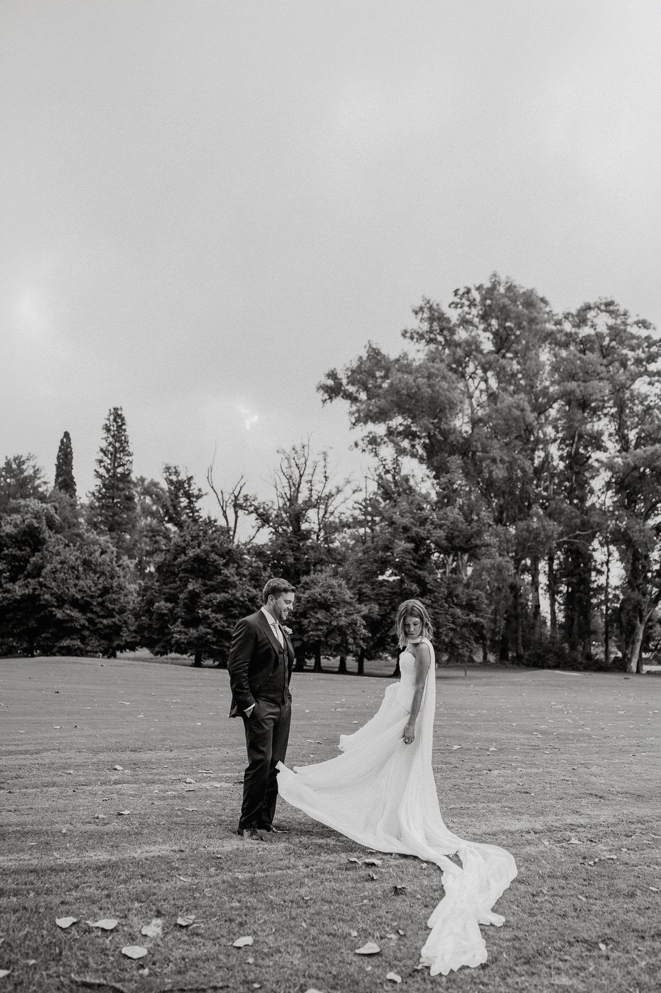 Black and white photo of bride and groom standing in open field, wedding dress flowing in the wind, trees in background.