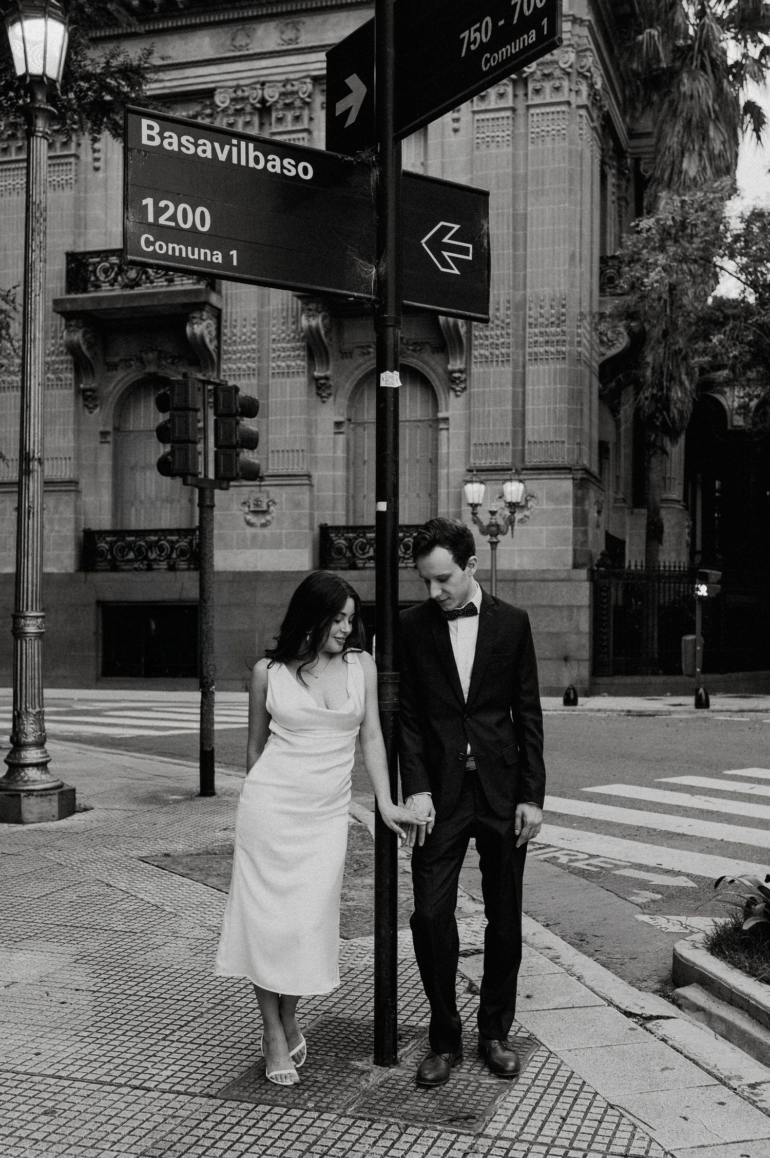 A couple in formal attire stand together under a Basavilbaso street sign in a black and white urban setting.