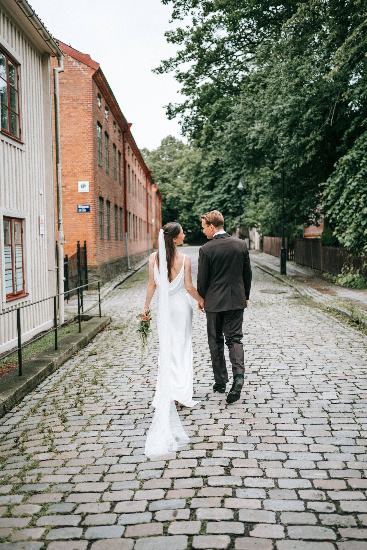 brudpar som går hand i hand längst kullerstensgator i göteborg nära röda sten och sockerbruket innan vigsel i st birgittas kapell, fotograf