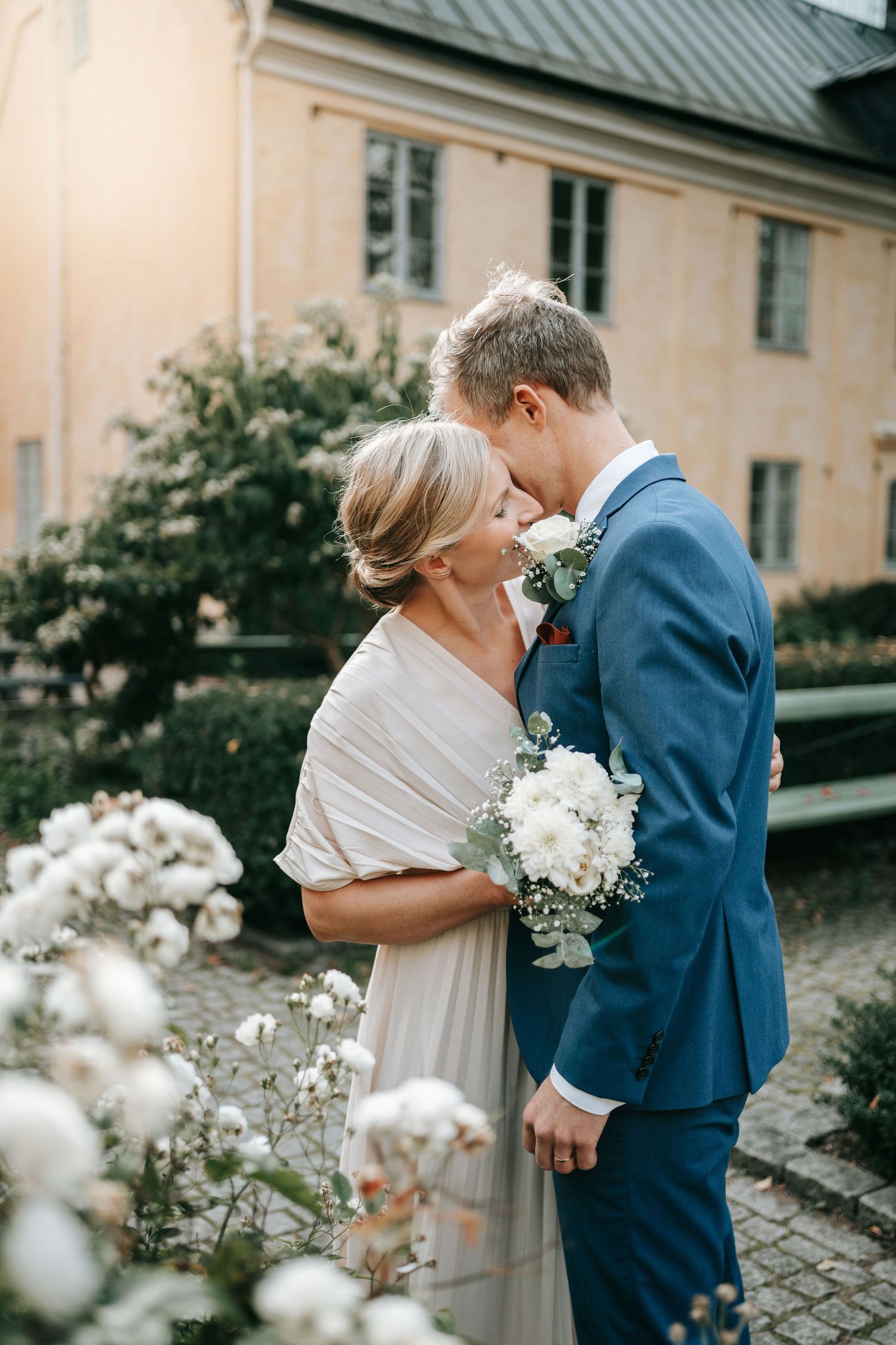Brud och brudgum kysser varandra i parken vid kronhusbodarna i göteborg innan borglig rådhusvigsel omgivna av vita blommor, bröllopsfotograf