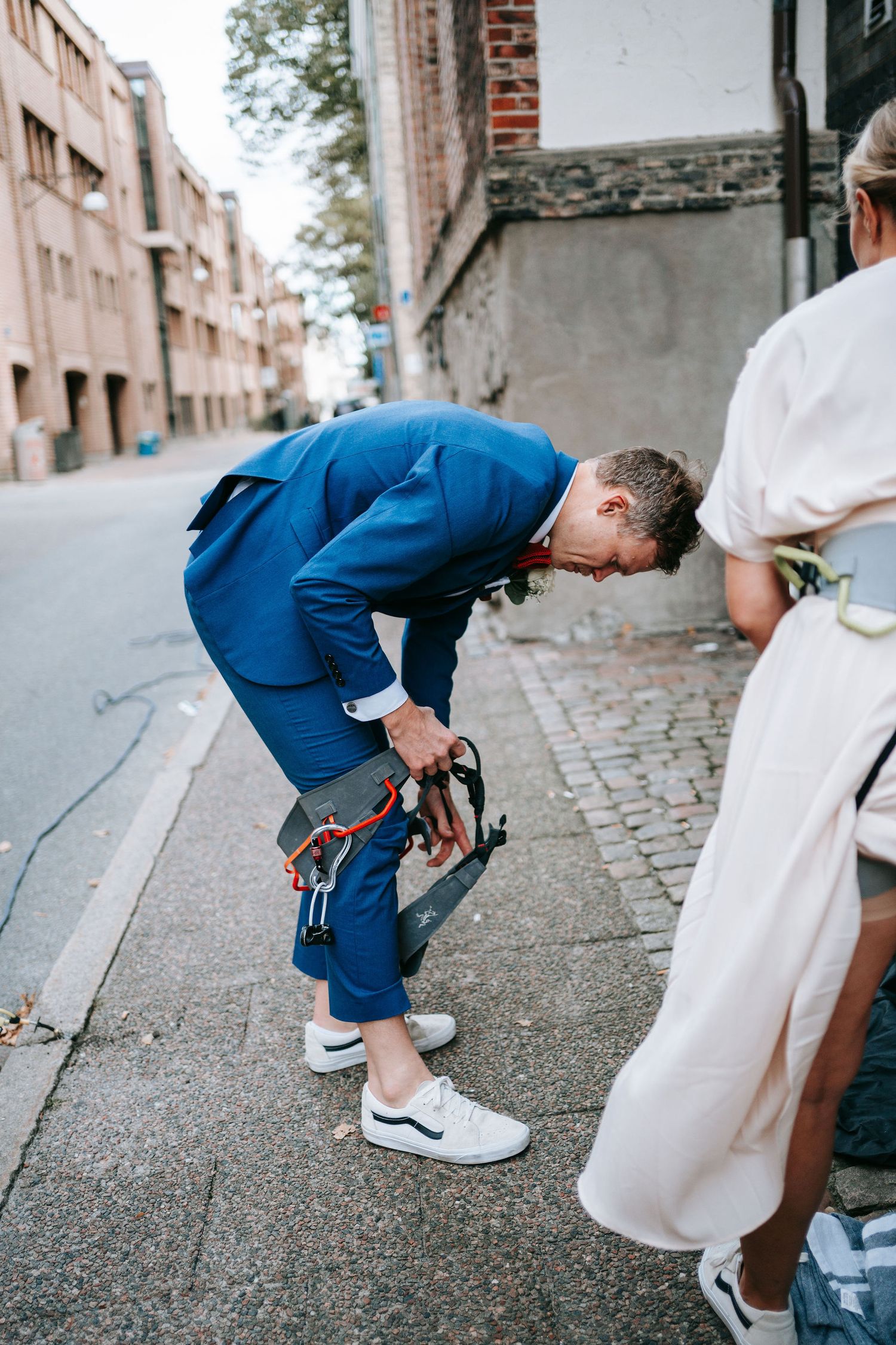Brudgum förbereder klätterutrustning på gatan medan bruden står bredvid i stadsmiljö i Göteborg, bröllopsfotografering