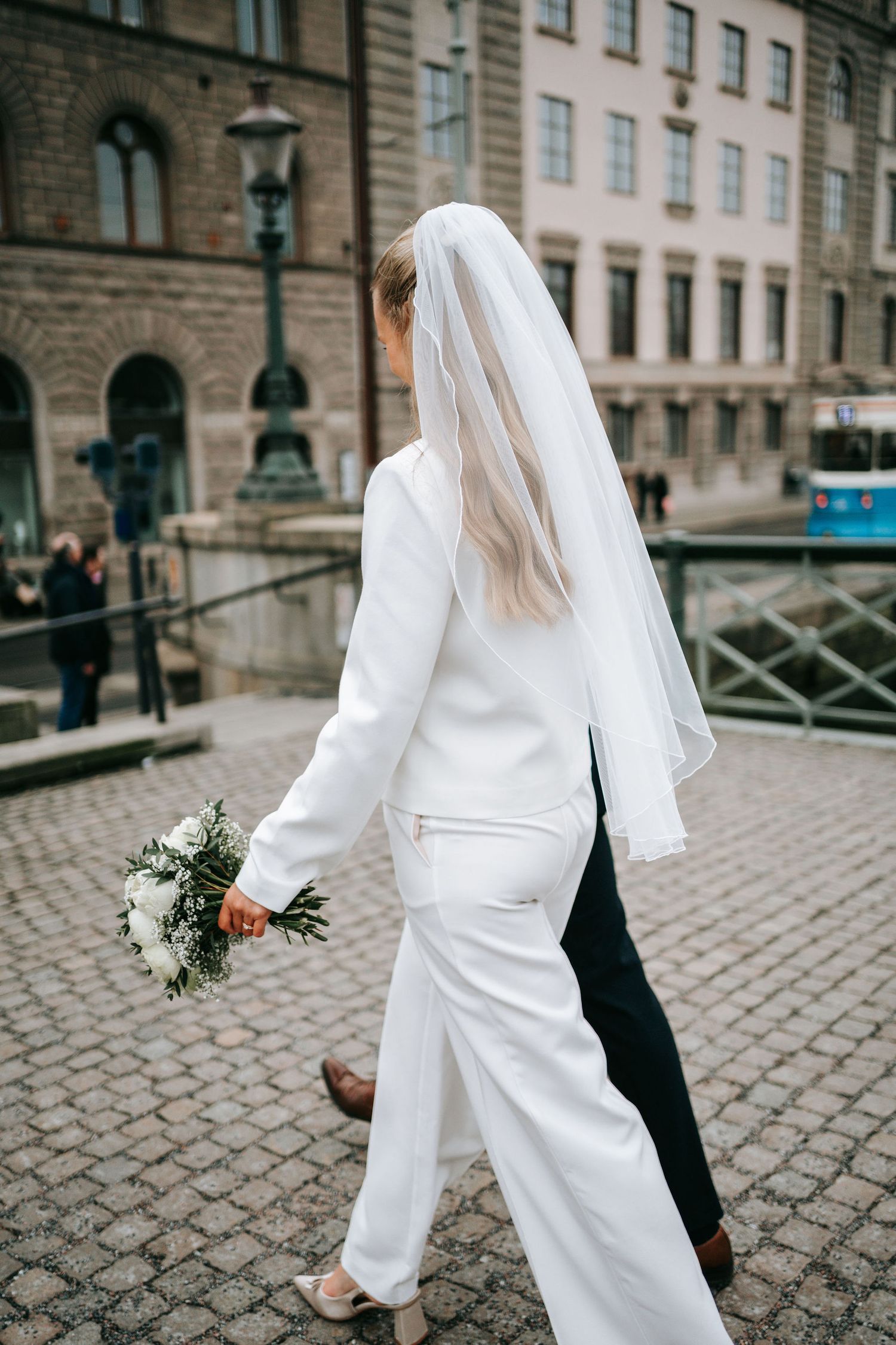 Brud i vit kostym och slöja promenerar på kullerstensgata vid rådhuset i Göteborg med brudbukett i handen. bröllopsfotografering göteborg