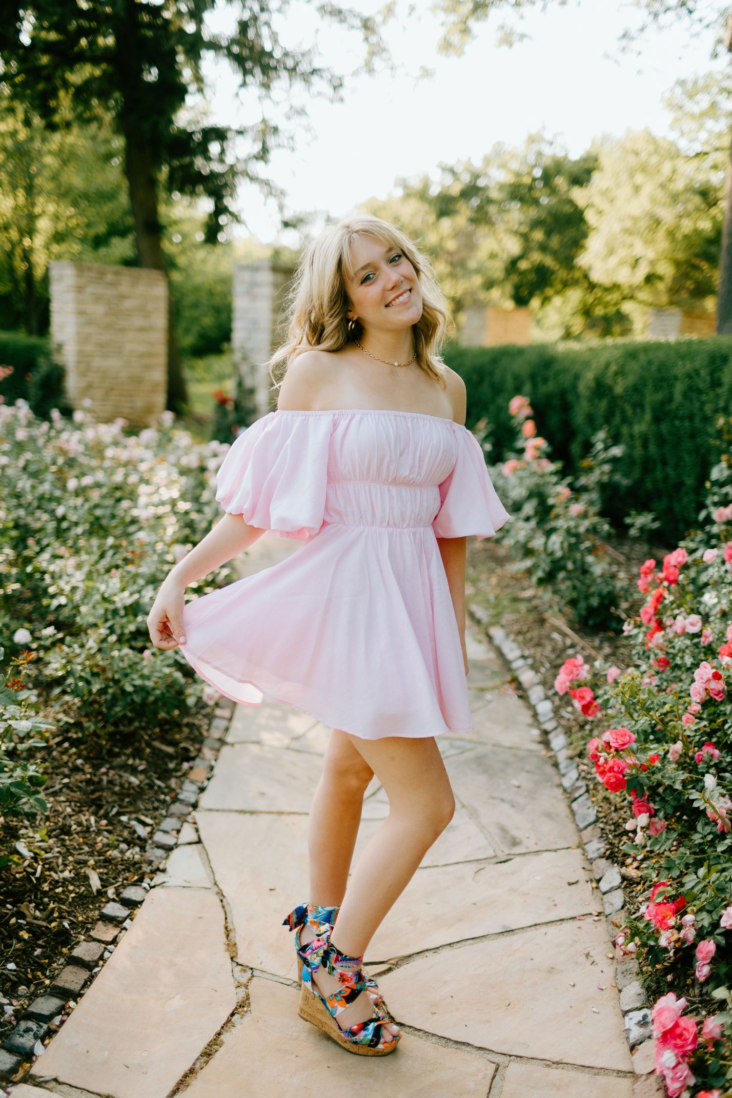 Young woman in pink off-shoulder mini dress posing on garden path surrounded by blooming roses.