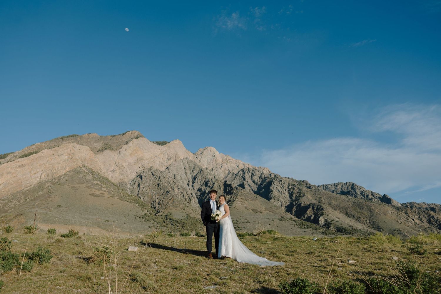 Bride and groom stand together with stunning mountain backdrop under clear blue sky.