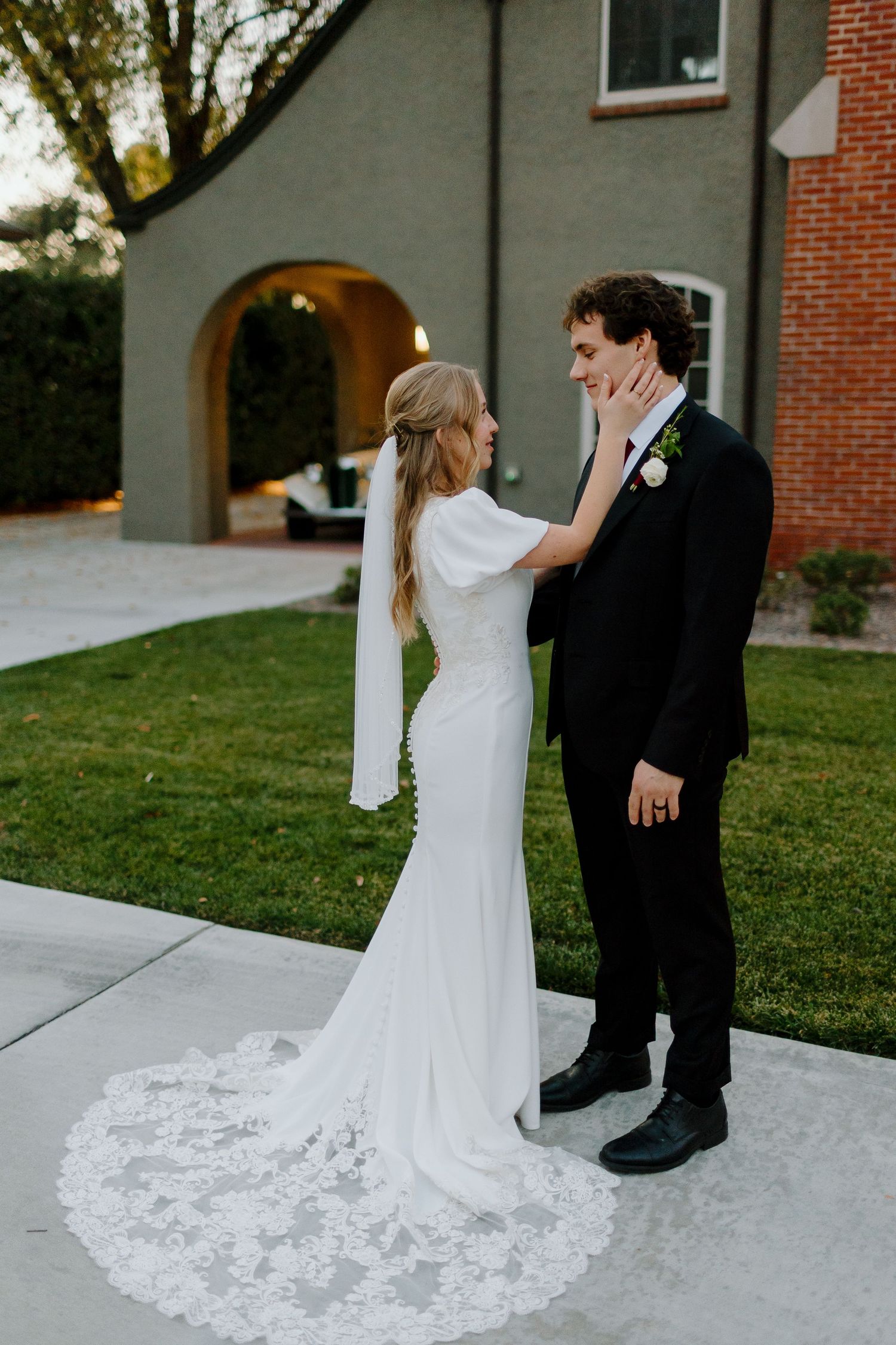 Bride in lace-train white gown tenderly touches grooms face outside historic brick building at dusk.