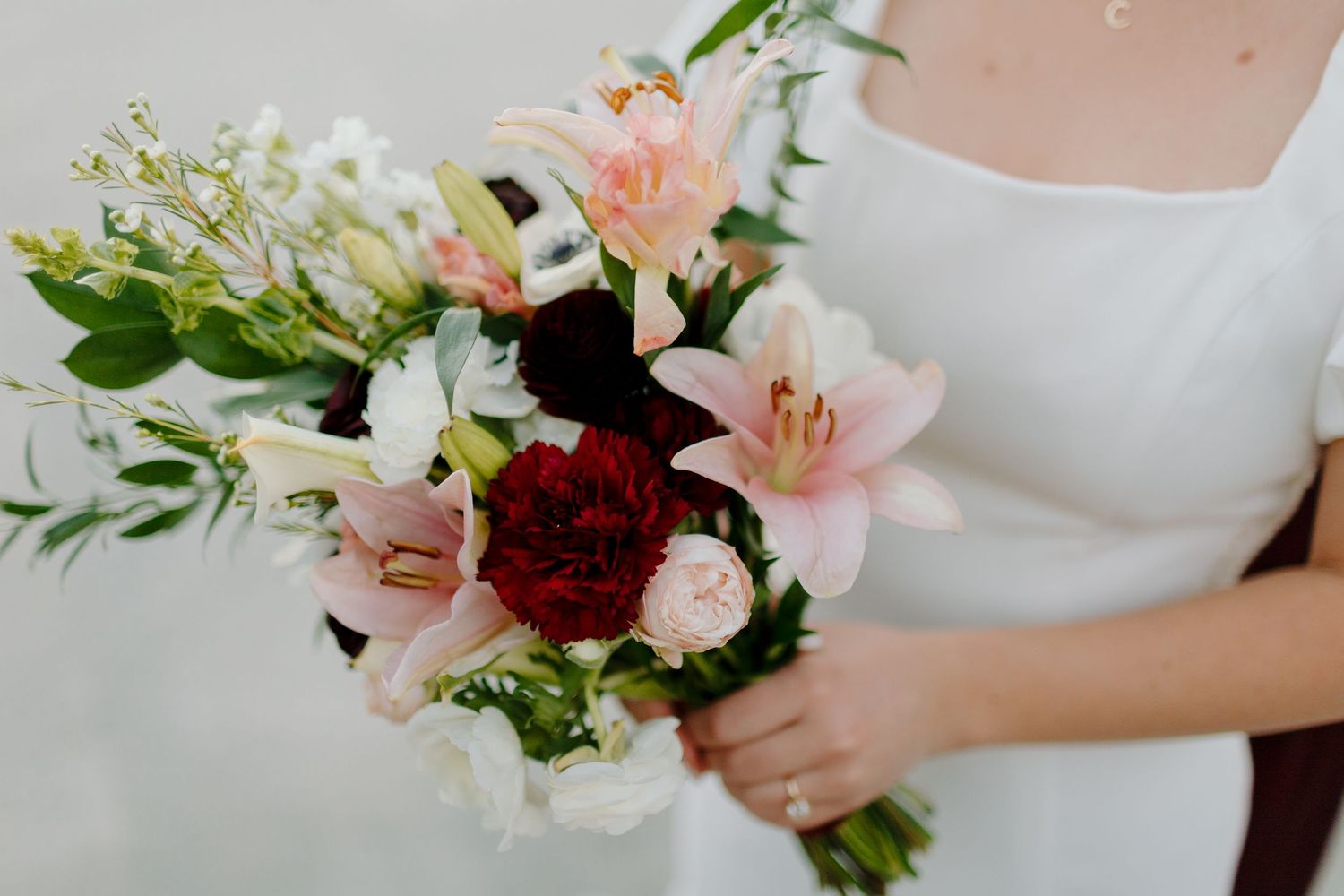 Bridal bouquet featuring pink lilies, burgundy carnations, and white flowers with green foliage.