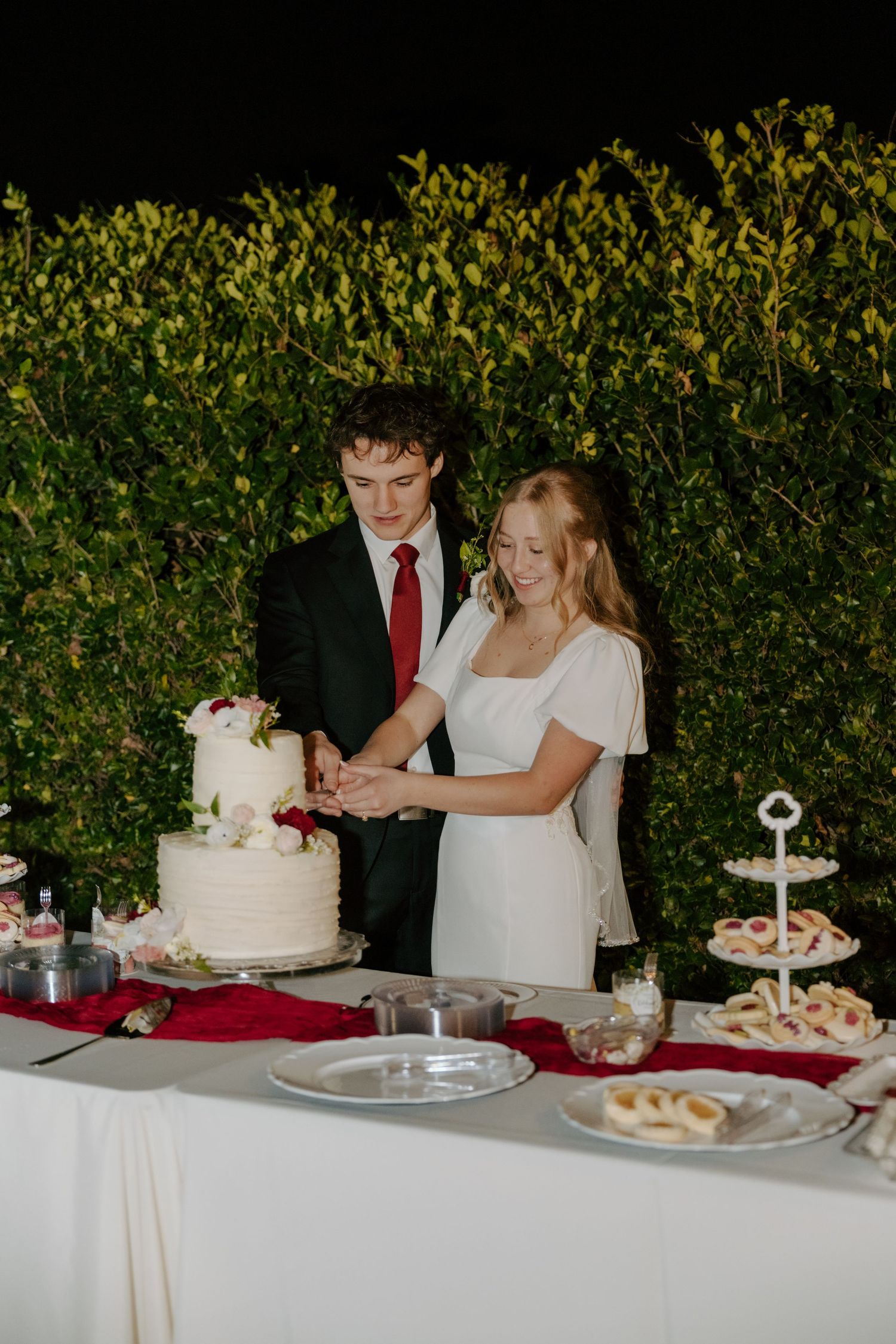 Bride and groom cut white floral wedding cake at reception table against green ivy backdrop.