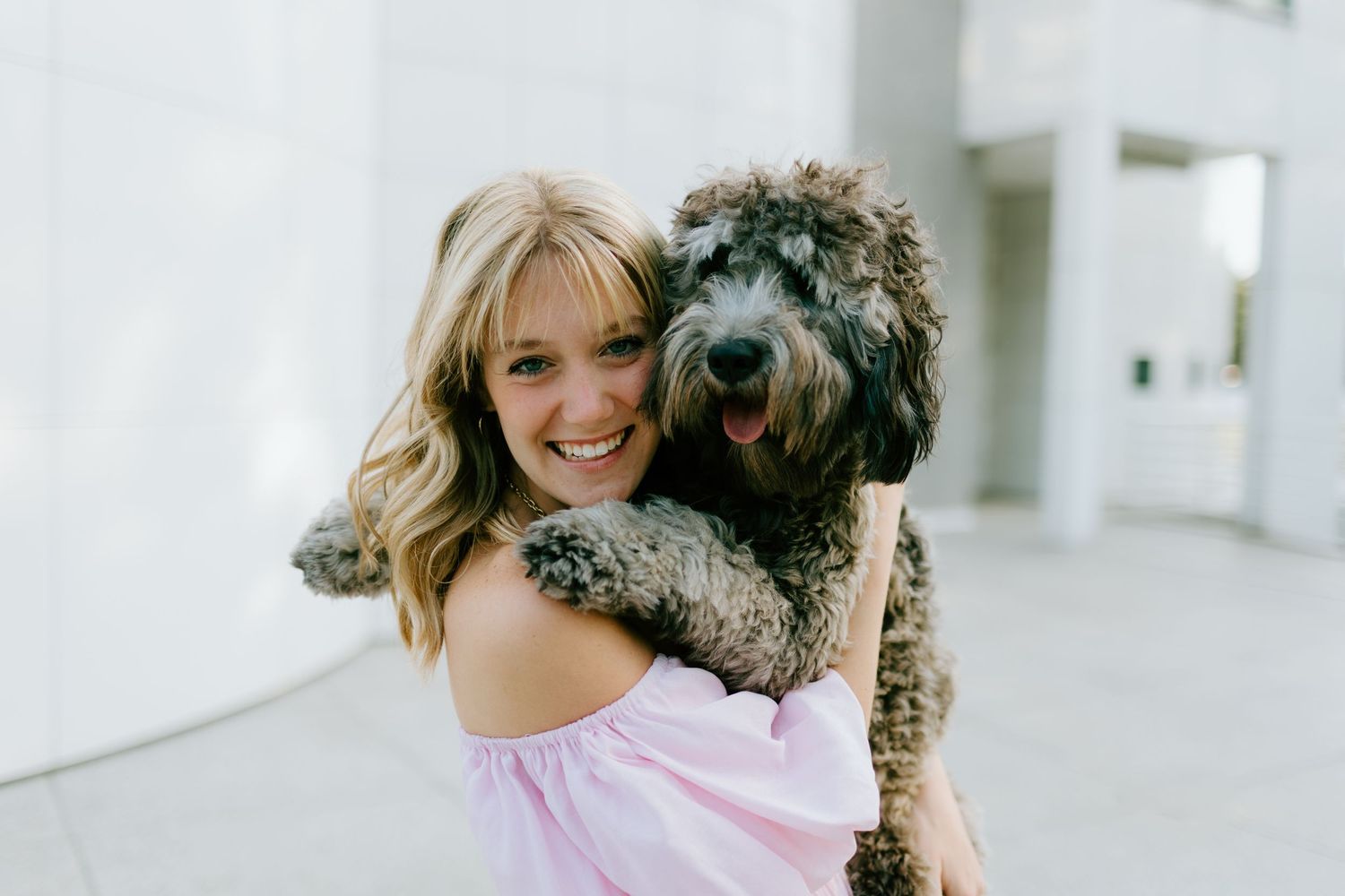 Smiling young woman in pink off-shoulder top hugging a fluffy brown doodle dog outdoors.