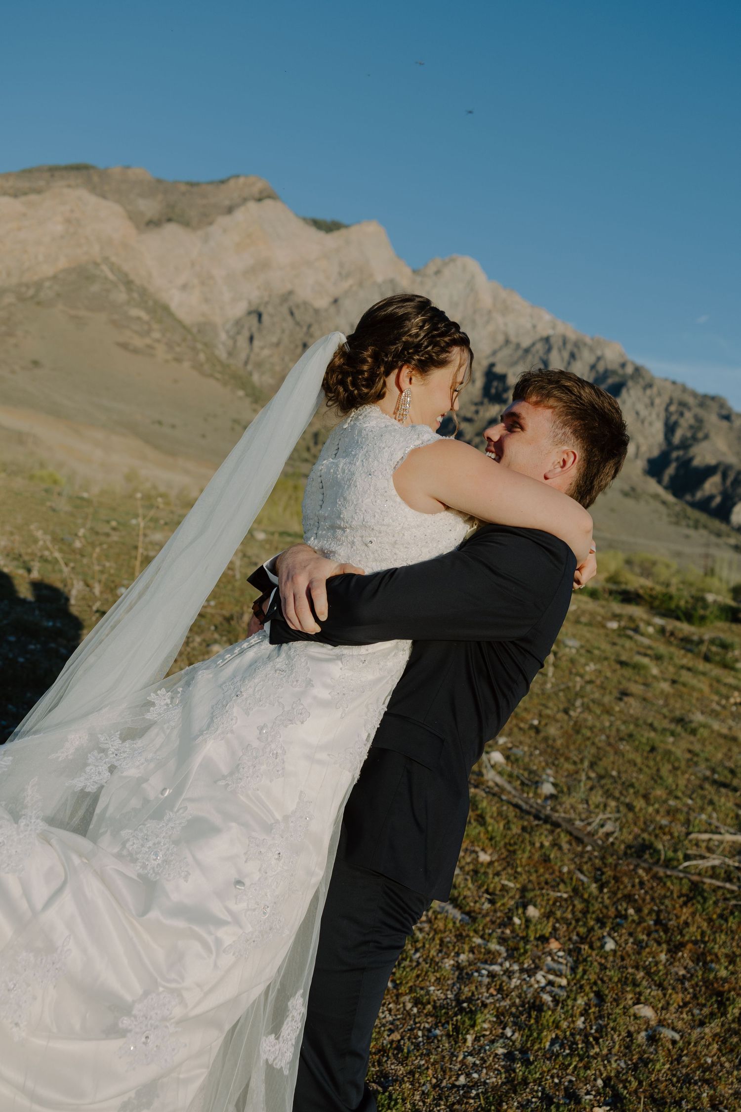 Groom lifting bride in white lace gown and veil against stunning mountain landscape on wedding day.
