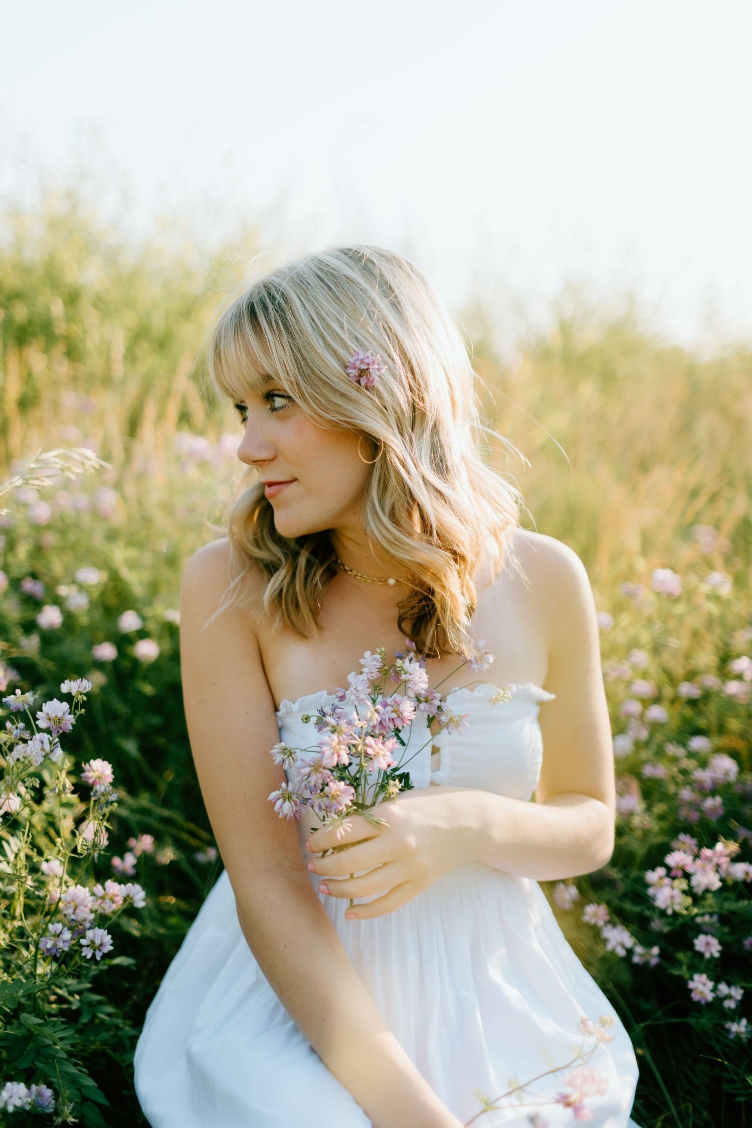 Blonde woman in white strapless dress holding pink wildflowers while sitting in a sunlit meadow of blooming flowers.