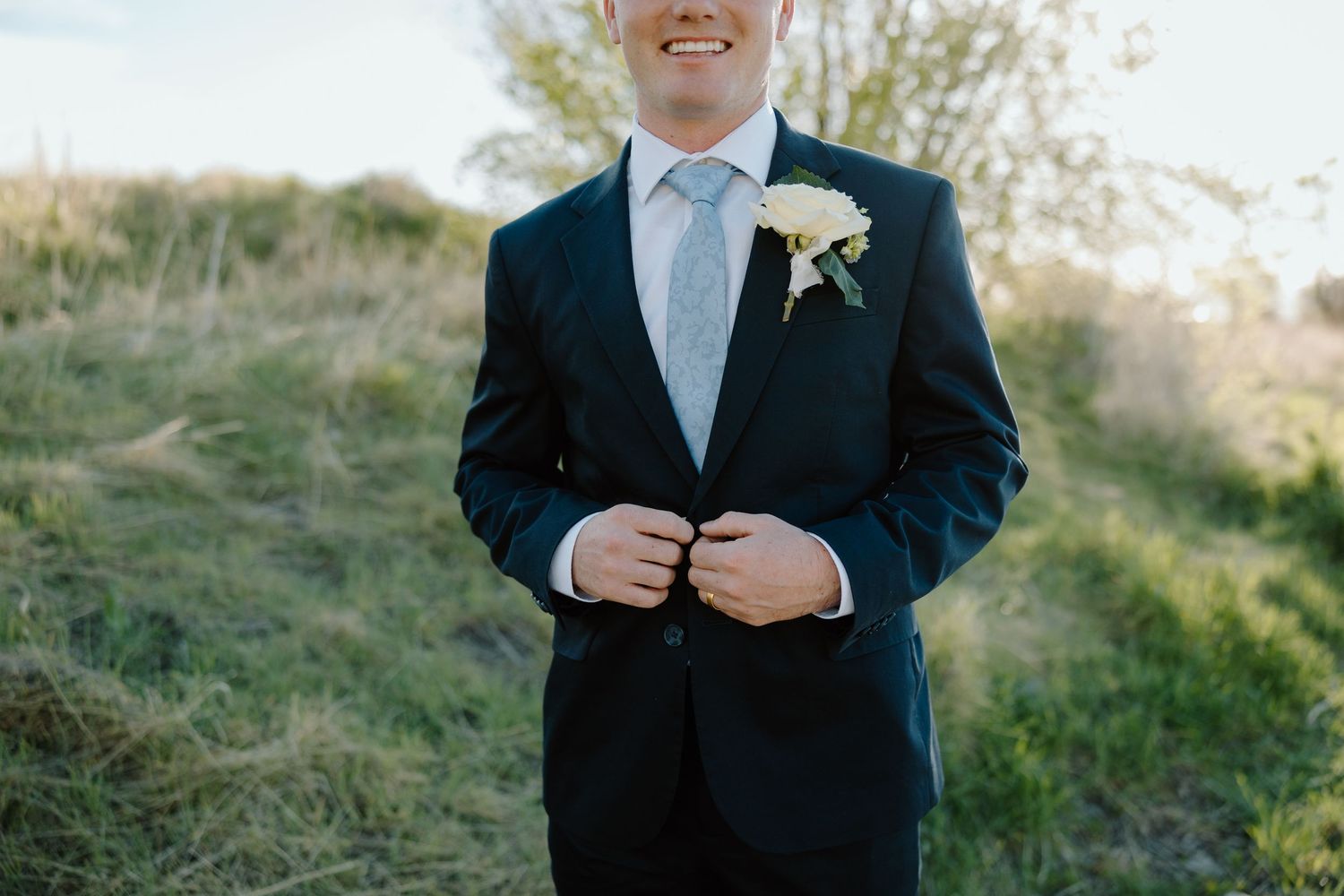Smiling groom in dark navy suit with white boutonniere buttoning jacket outdoors in green field.