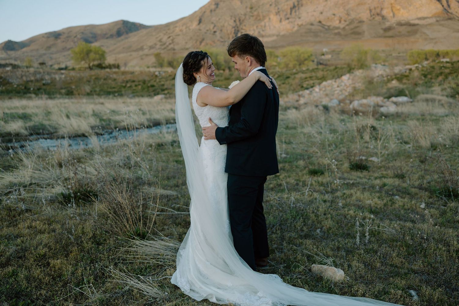 Bride and groom embrace in a scenic outdoor wedding portrait with mountains and green fields in the background.