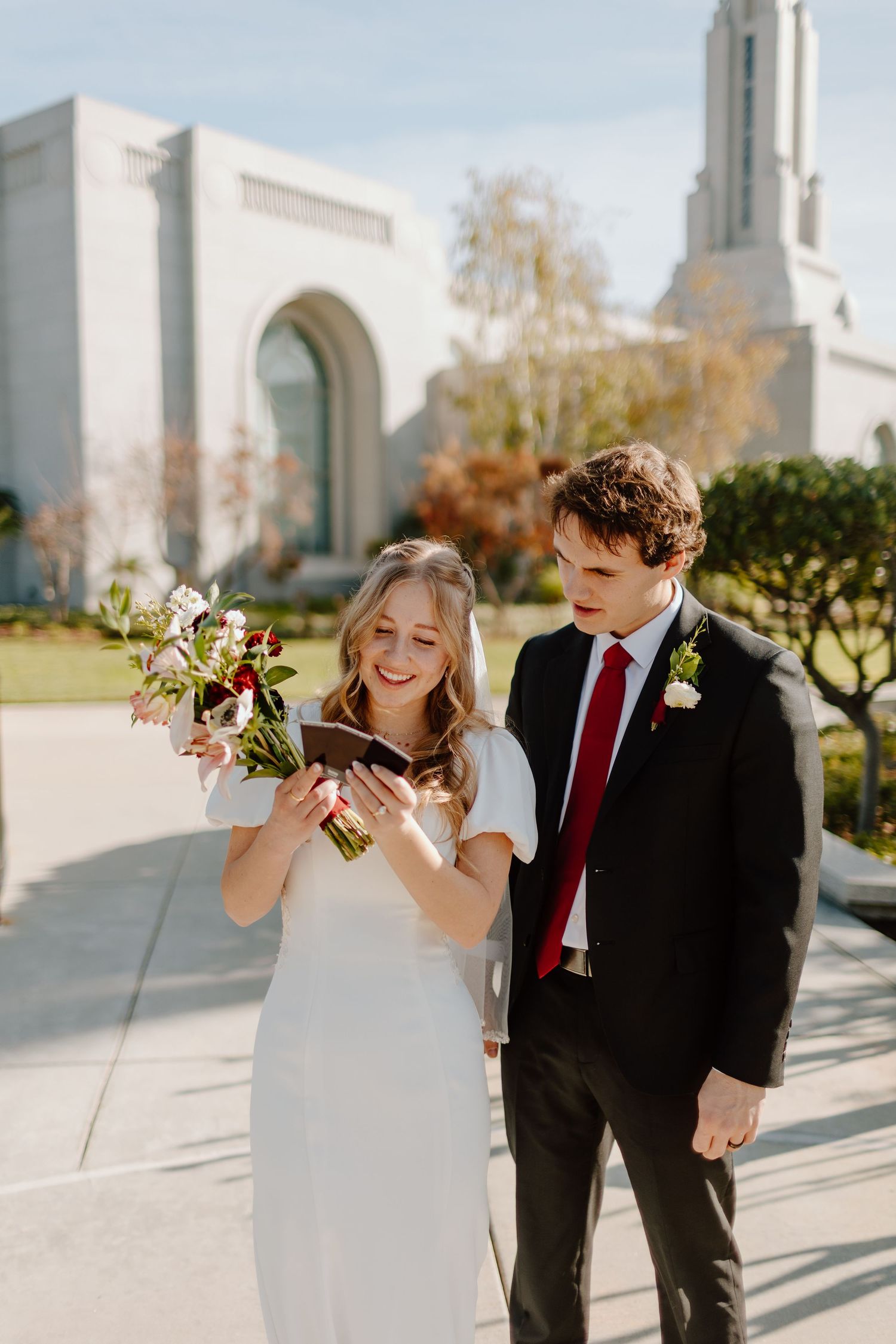 Bride and groom smiling at photos outside an LDS temple on their wedding day, bride holding a floral bouquet.
