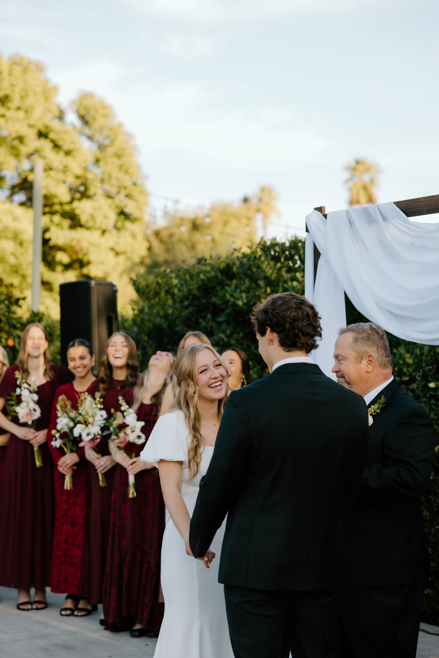 Joyful bride laughs while holding groom's hands at outdoor wedding ceremony with bridesmaids in burgundy dresses.