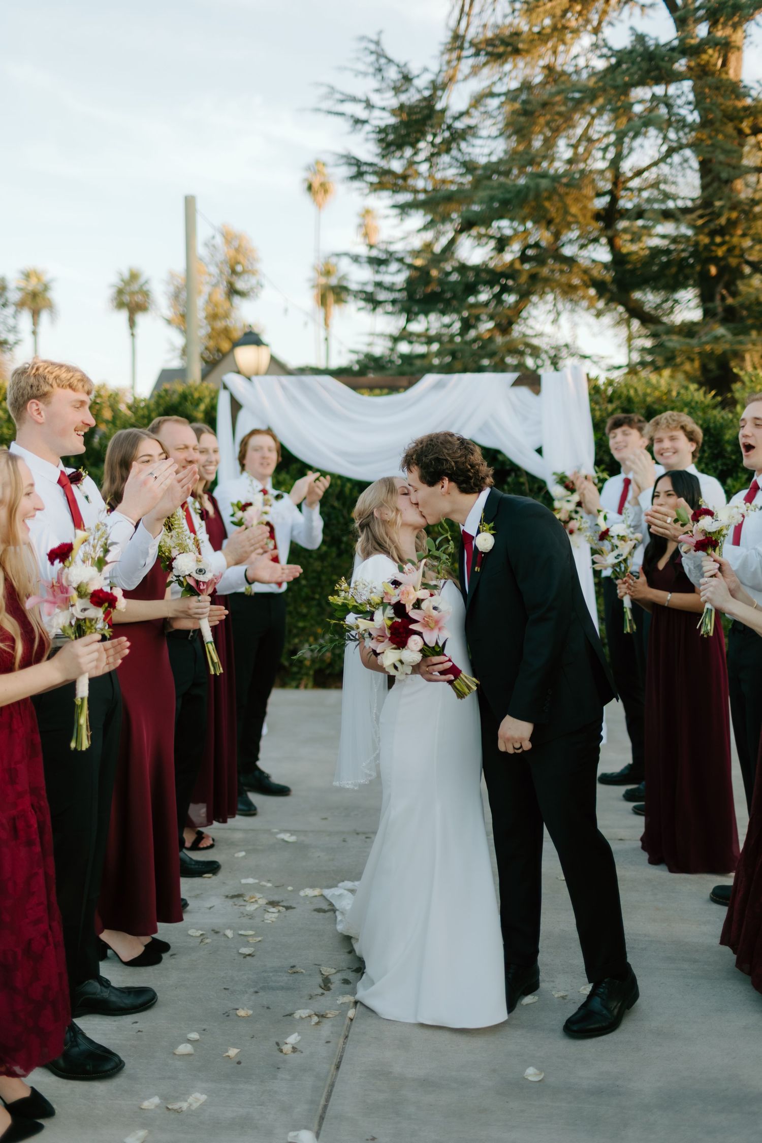 Newlyweds share a first kiss while wedding party in burgundy attire cheers along the aisle outdoors.