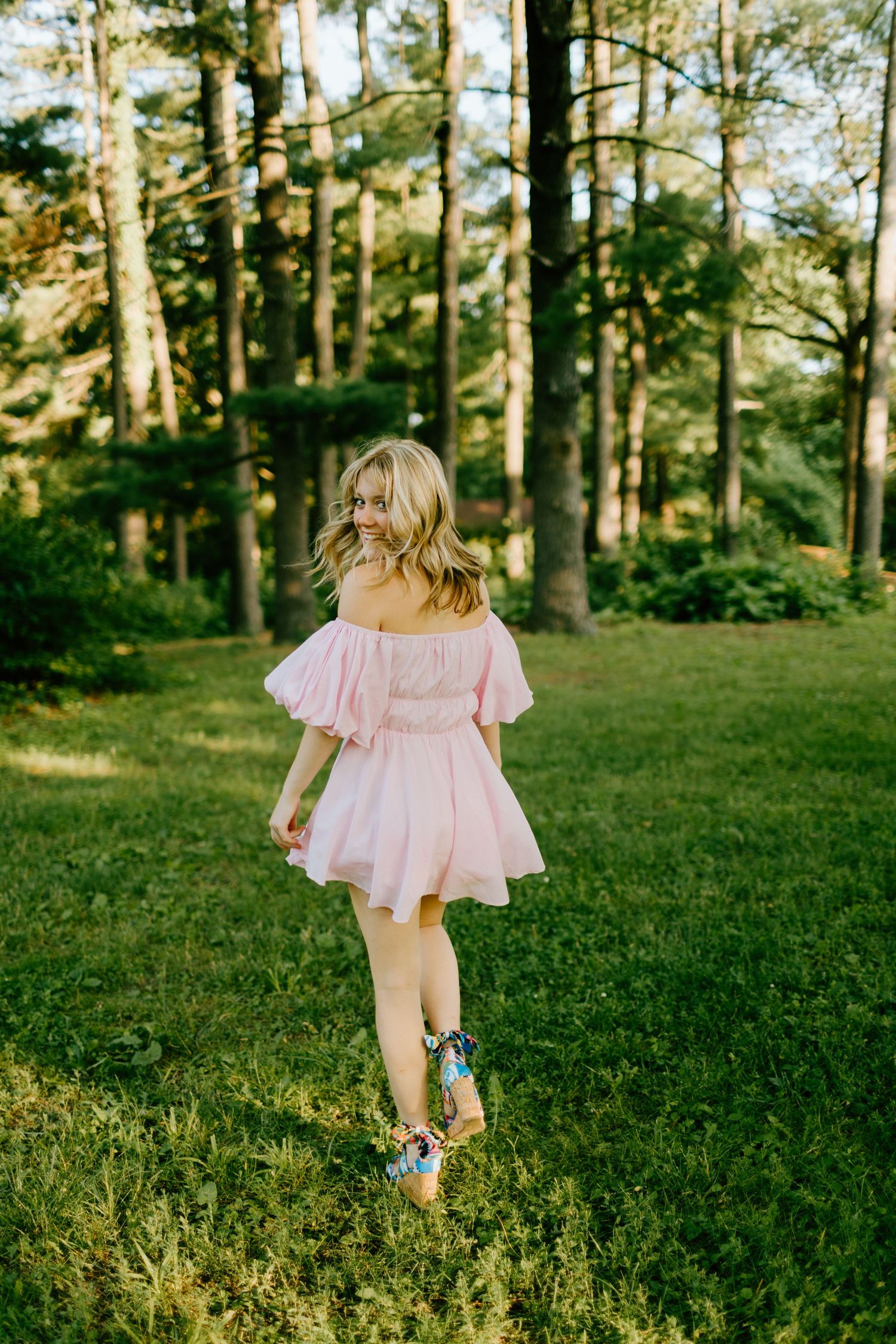 Young woman in pink off-shoulder mini dress walking on green grass among tall trees in golden hour light.