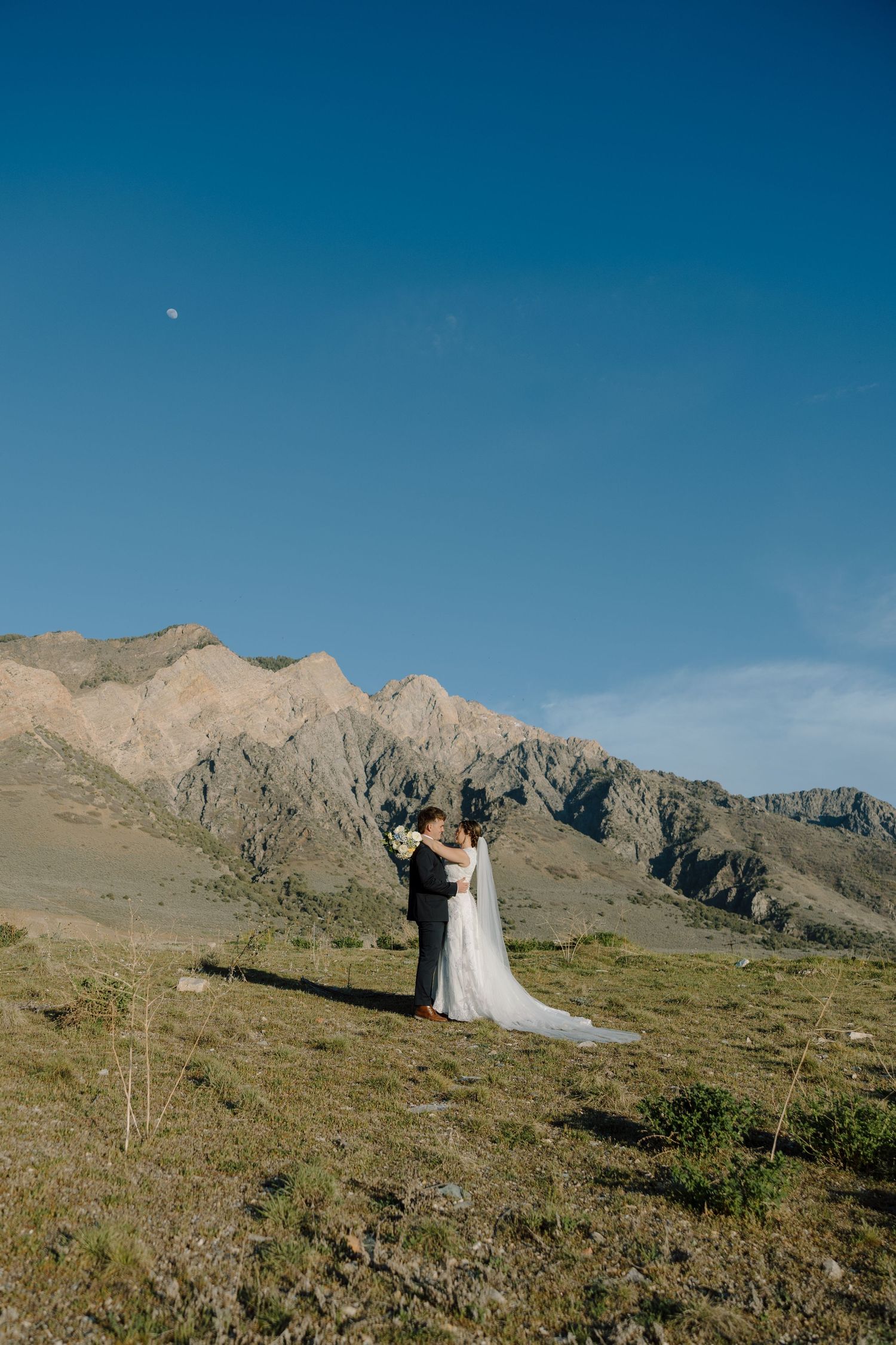 Bride and groom embrace before dramatic mountain backdrop under a clear blue sky.