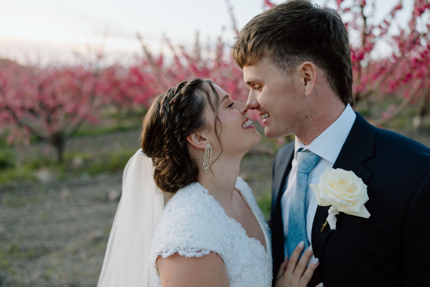 Happy newlyweds smile intimately together with pink cherry blossom trees glowing behind them.