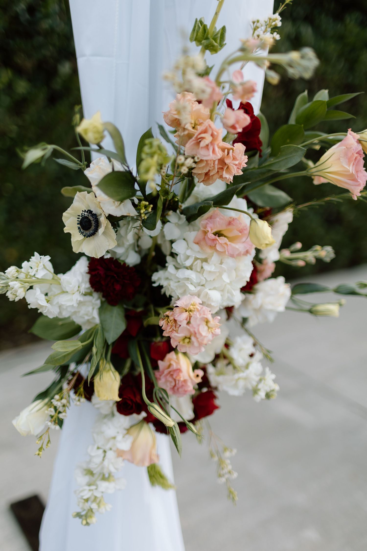 Elegant wedding arch floral arrangement with white, pink, and burgundy blooms, anemones, and lush greenery on white draping.