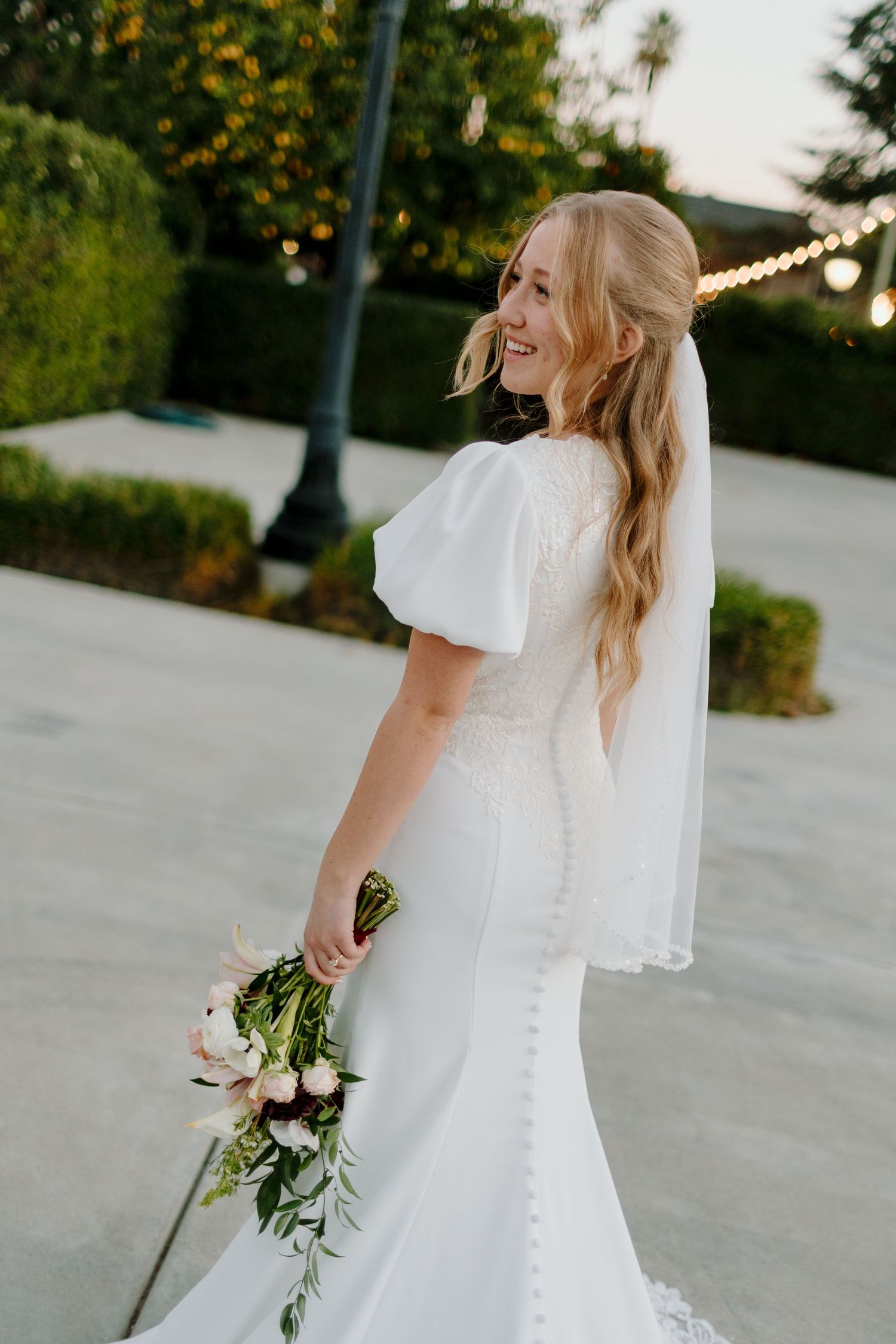 Smiling bride in elegant white gown with puff sleeves and veil holding a bouquet of pink and green flowers.